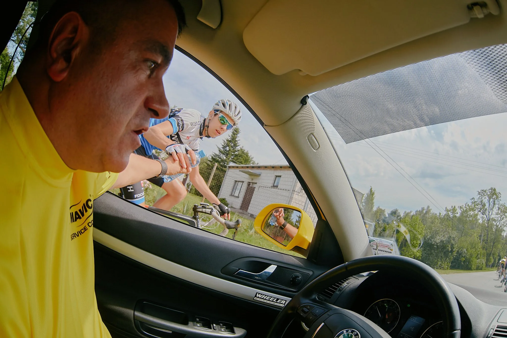 A cyclist in white and blue gear and sunglasses leans forward on their bike, seen through the open window of a yellow car. Two other cyclists are riding on the road behind them, and there's a white house with a fence and trees visible in the backgrou