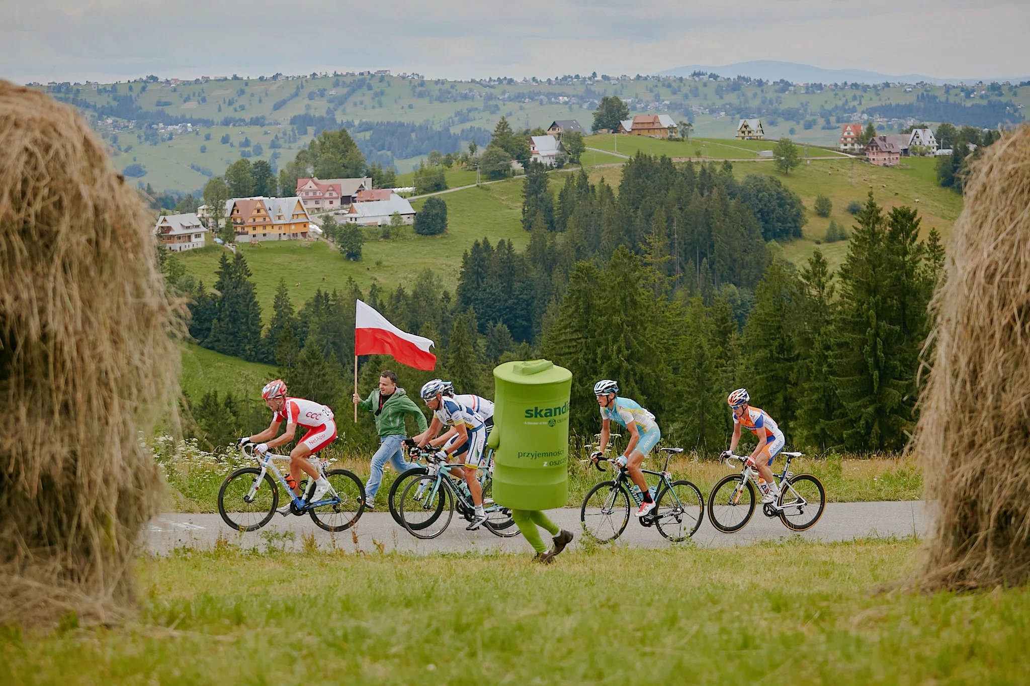 Cyclists participating in a race or event through a scenic green countryside with houses on rolling hills in the background, viewed through hay bales