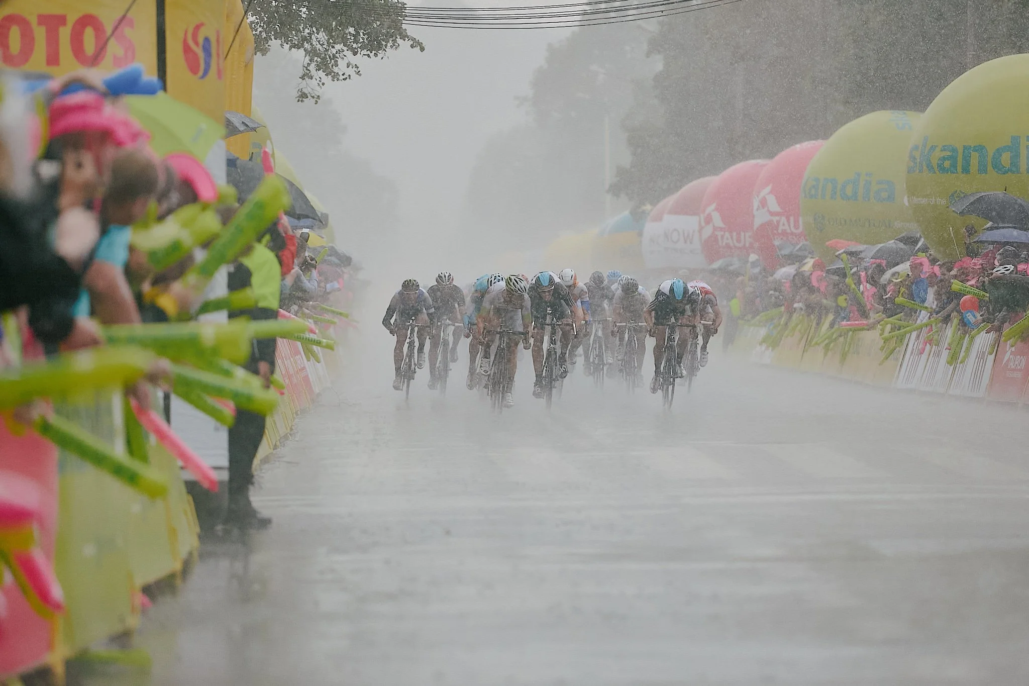 Cyclists racing in heavy rain during a professional cycling race with spectators holding umbrellas and balloons lining the course.