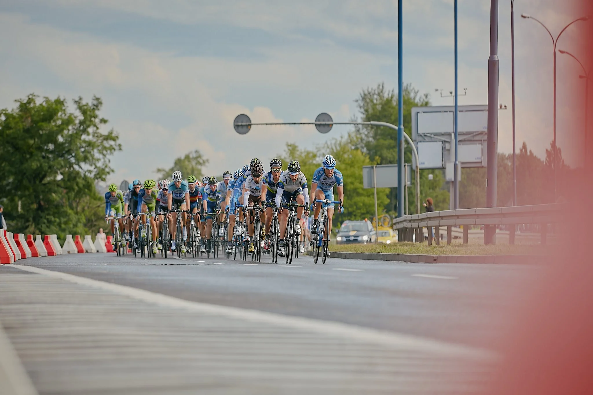 A large group of cyclists riding on a city road during daytime, with some green trees and blue sky in the background.