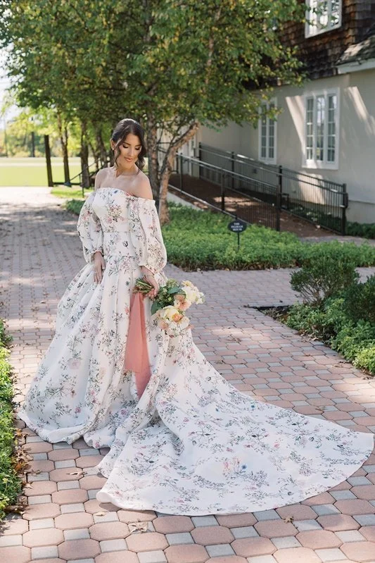 A bride in a floral wedding gown holding a bouquet standing on a brick sidewalk near greenery and a white building.