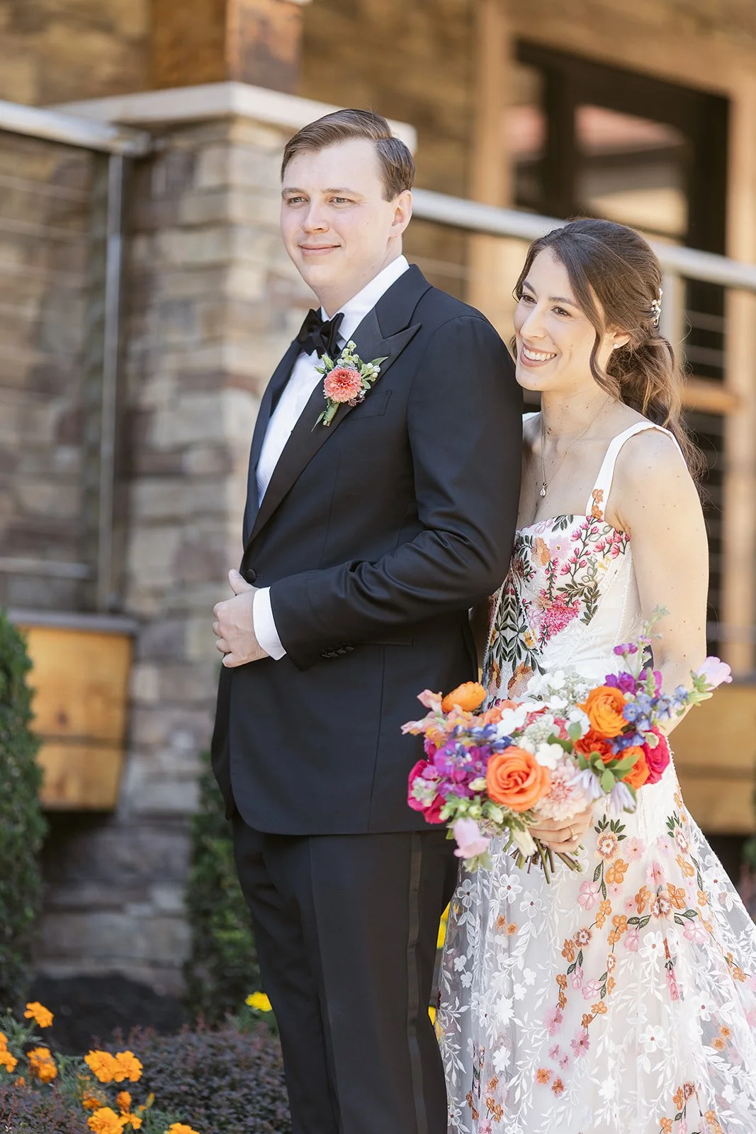 A bride and groom standing outdoors during their wedding. The groom is wearing a black tuxedo with a white shirt and a black bow tie, while the bride is wearing a floral lace wedding dress and holding a colorful bouquet of flowers. They are smiling and standing in front of a brick building.