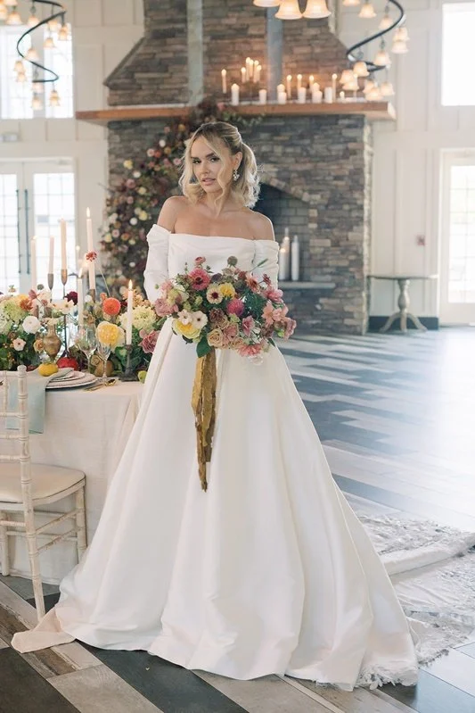 Bride in a white wedding gown holding a large bouquet of flowers, standing in a decorated reception hall with candles, flowers, and a stone fireplace.