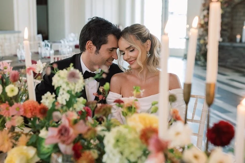 A happy bride and groom sitting close together at their wedding reception, surrounded by floral centerpieces and candles.