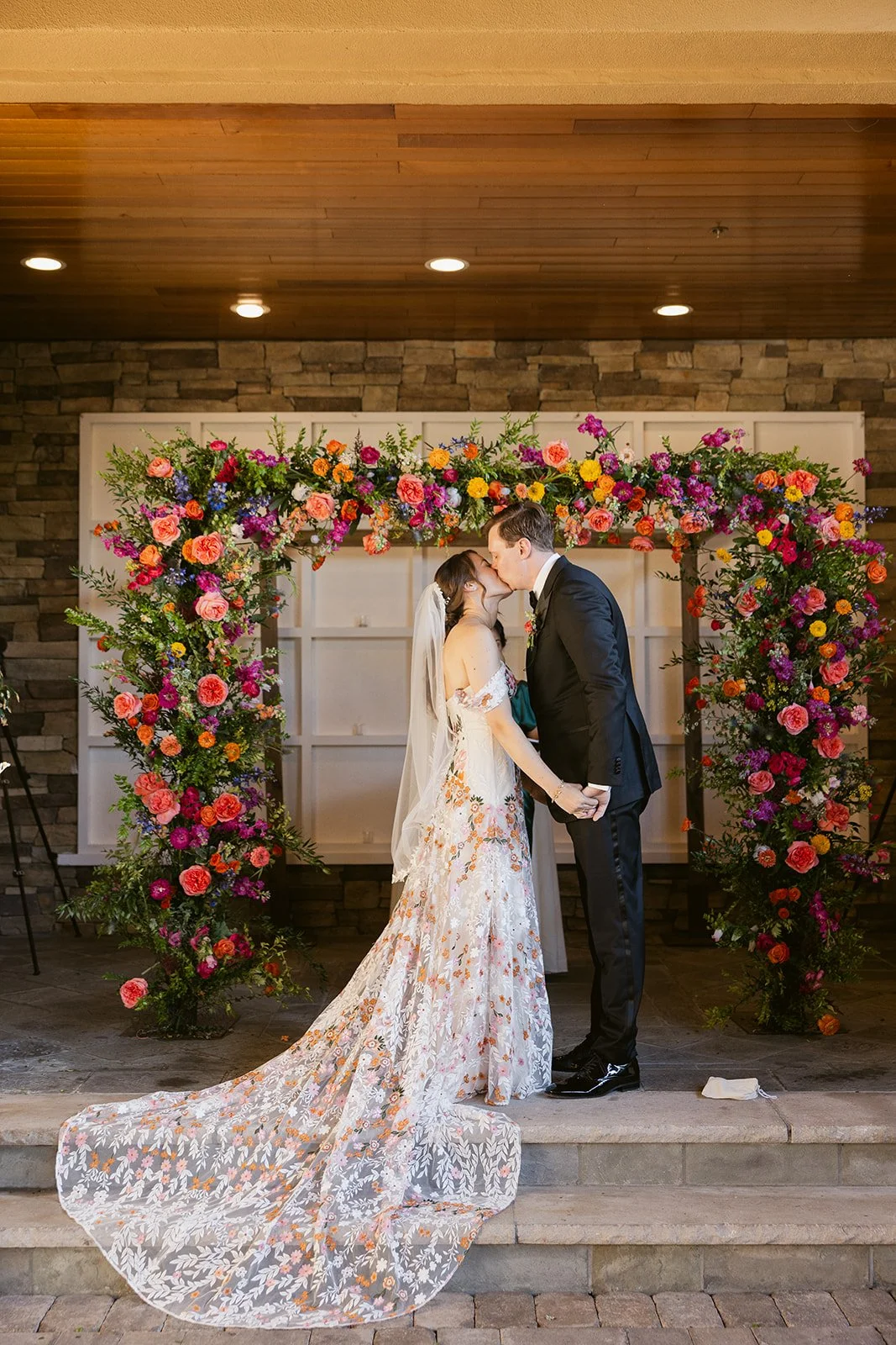 A bride and groom sharing a kiss during their wedding ceremony, standing under a vibrant floral arch.