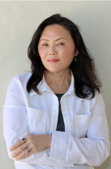 A woman with shoulder-length dark hair, wearing a white shirt over a gray top, standing in front of a light-colored wall with her arms crossed.