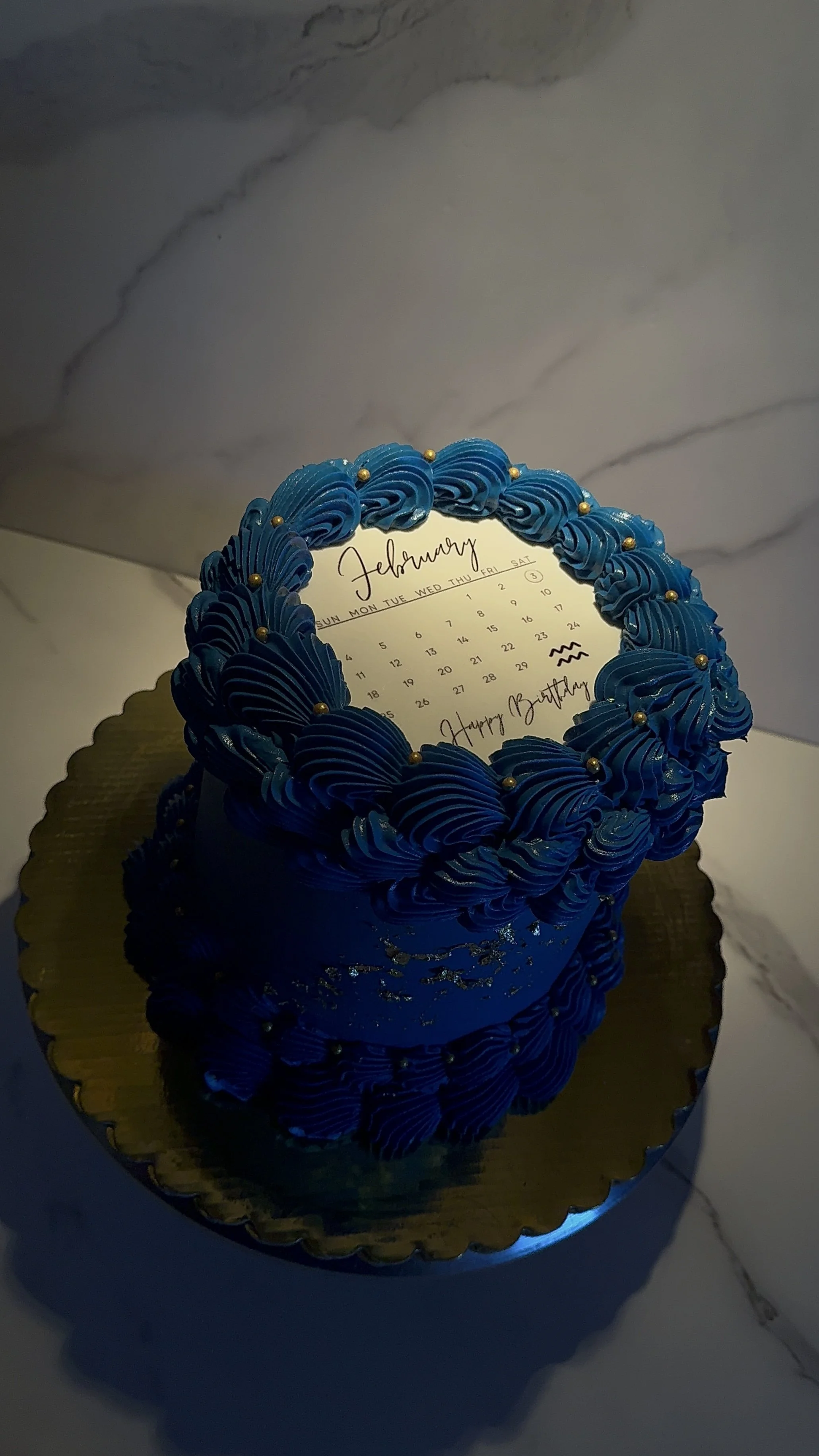 A round birthday cake with blue piping decorations, sitting on a gold cake board, with a calendar for February on top featuring the words 'Happy Birthday' and a small doodle.