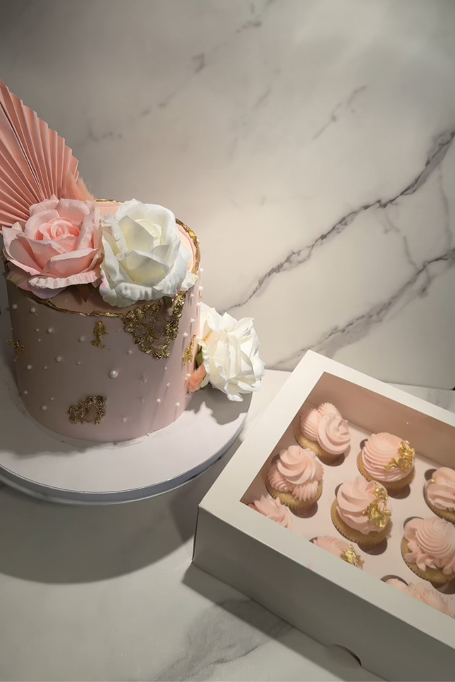 A pink cake decorated with white and pink flowers, gold accents, and a paper fan decoration. Next to it is a box of pink frosted cupcakes with gold decor, placed on a marble surface.