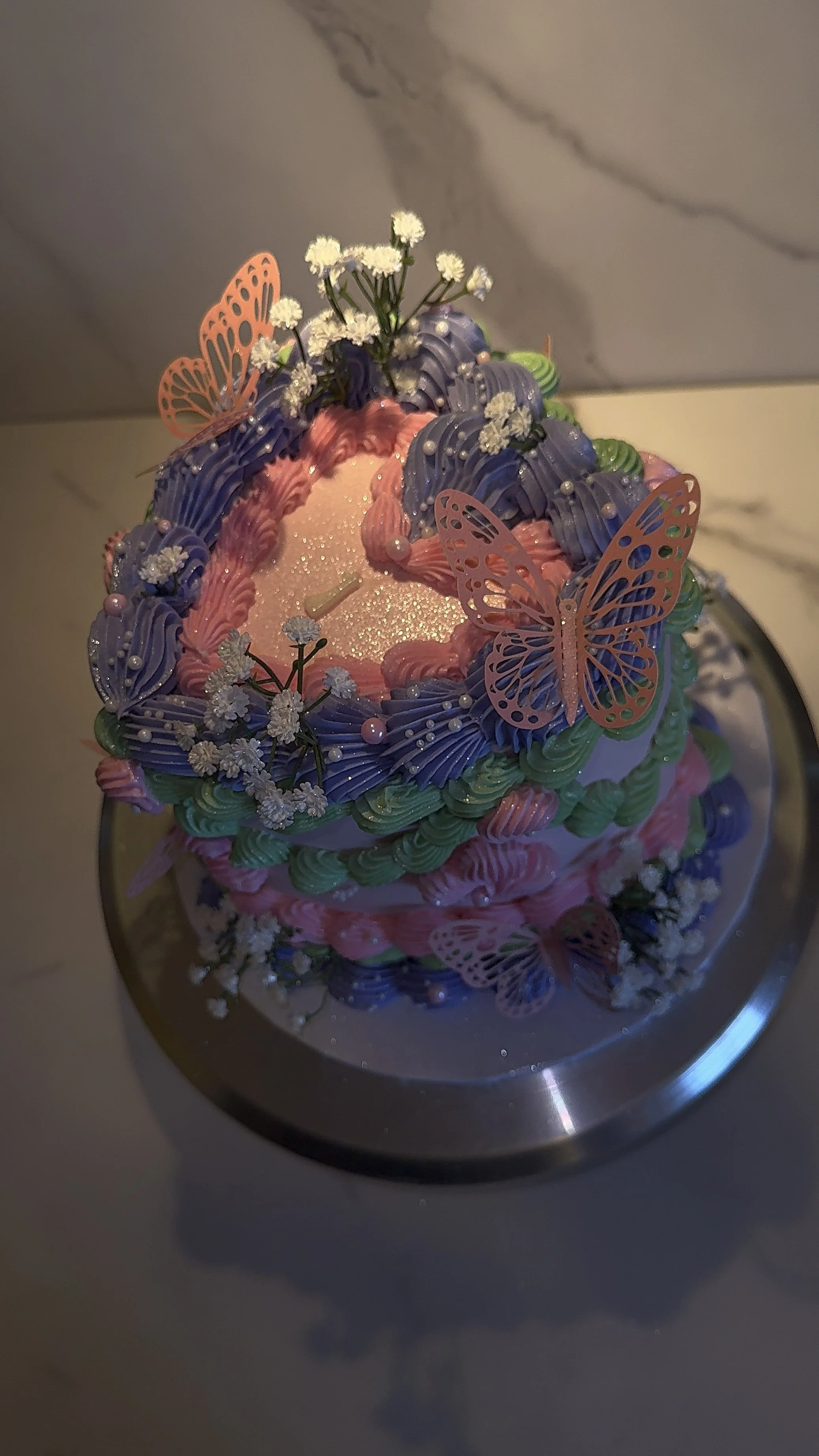 Decorative cake with pink, purple, and green frosting, adorned with small white flowers and orange butterfly decorations, placed on a reflective silver cake board.