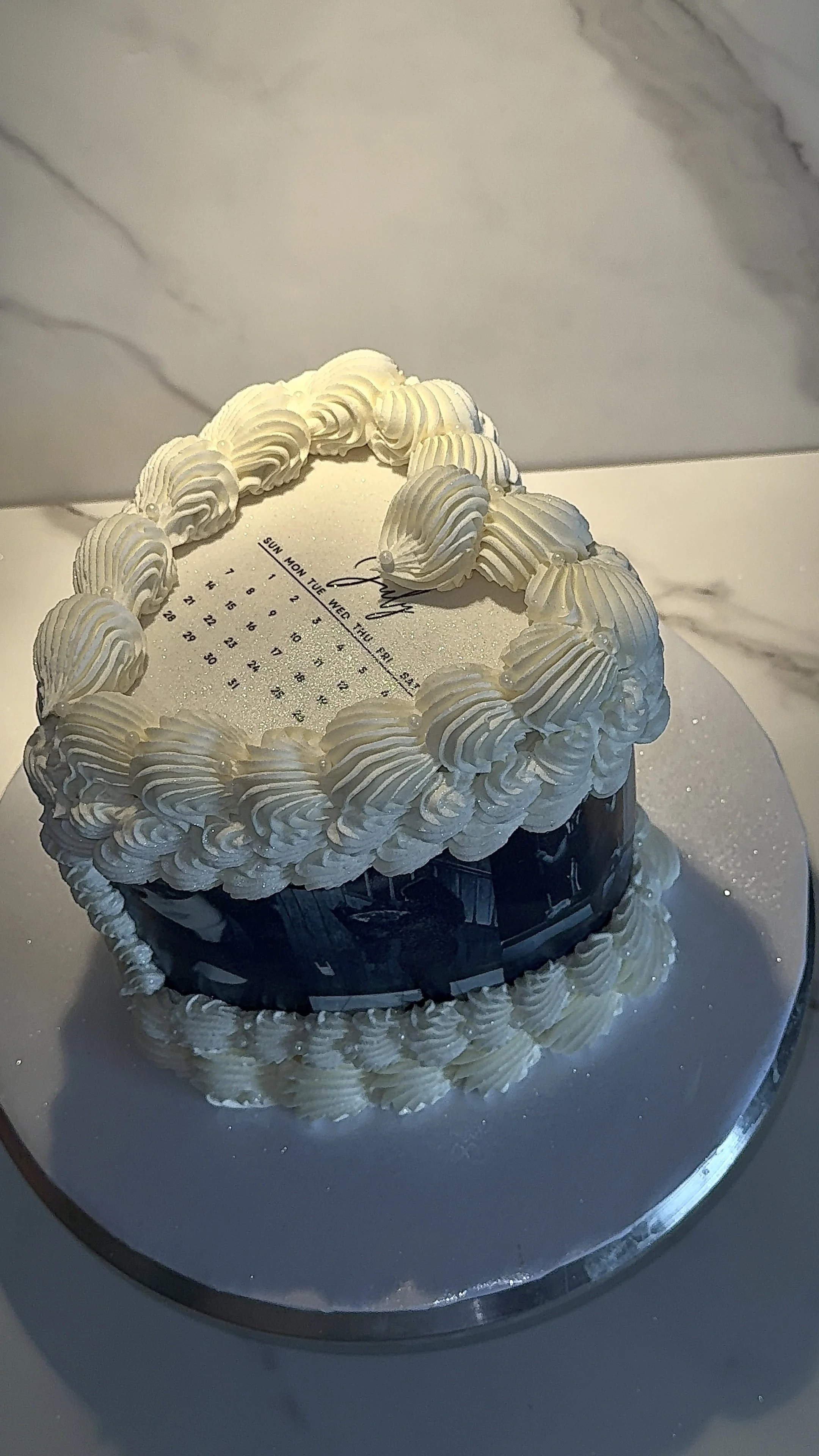 A round cake with white piped frosting decorations, reflecting on a mirrored cake stand, with a calendar and signature on the top surface.