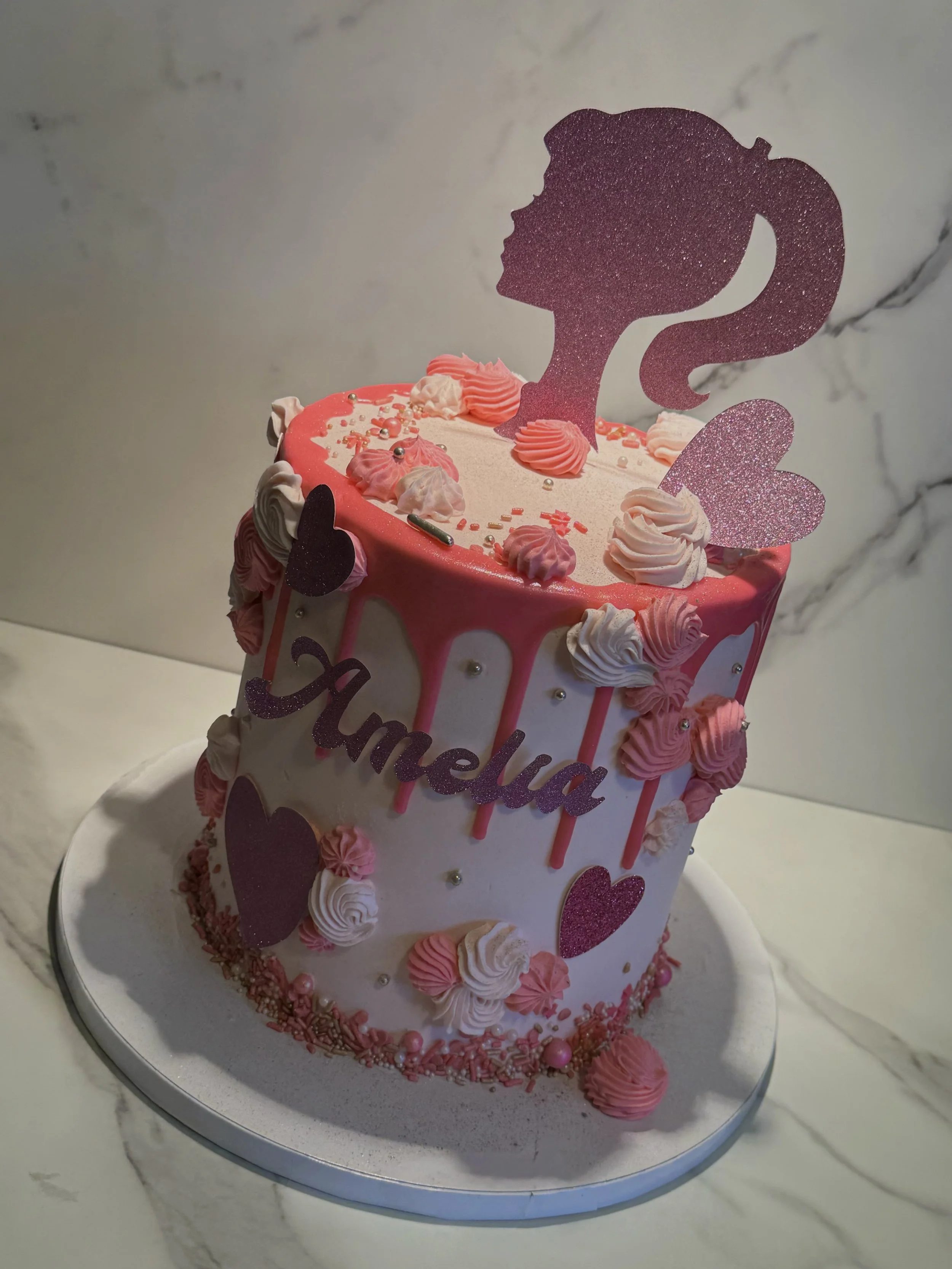 Pink and white birthday cake with a silhouette topper of a girl with a ponytail, decorated with pink and white frosting, hearts, sprinkles, and a 'Jameula' sign.
