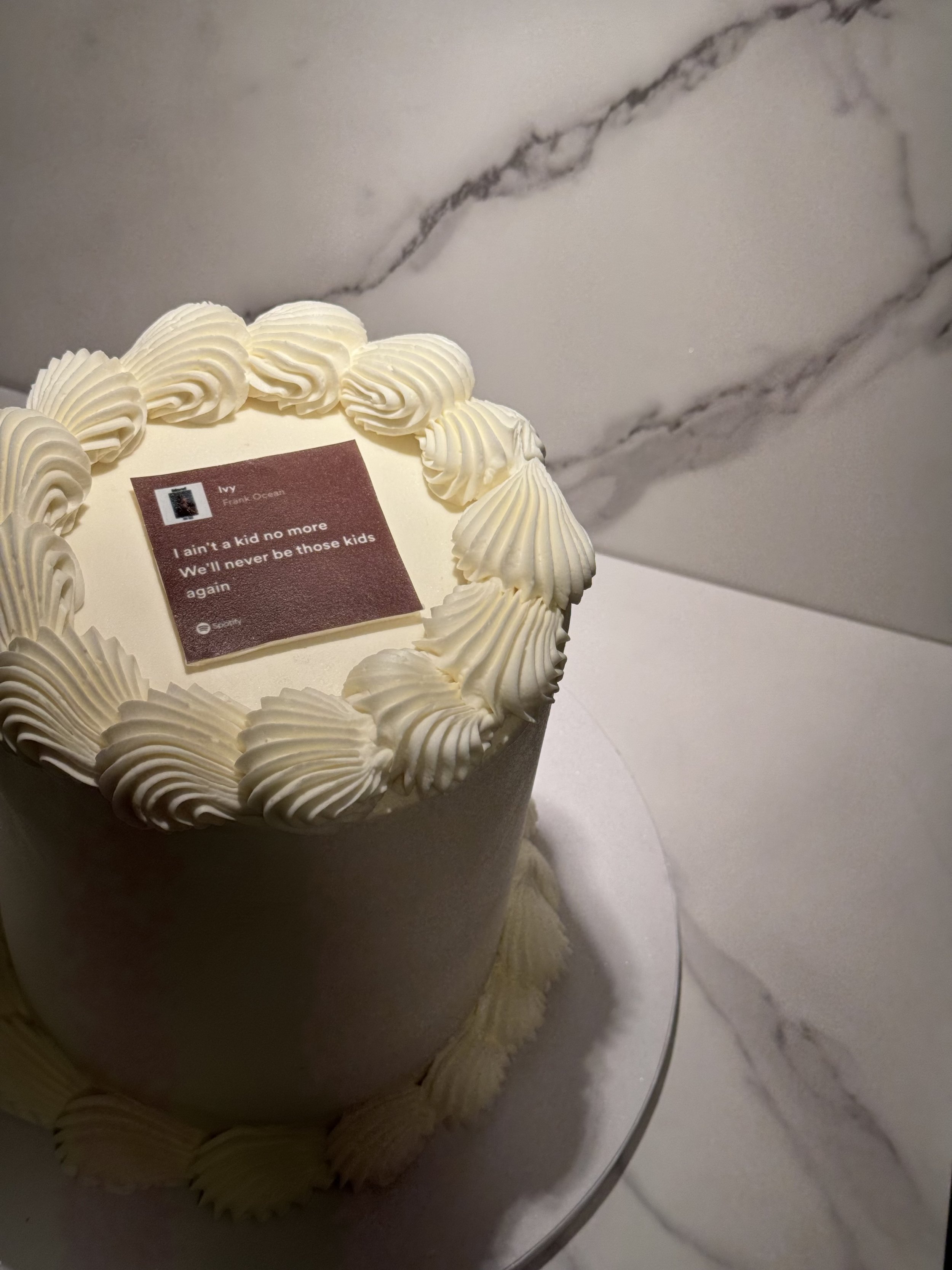 A white cake with piped cream decorations on top and around the base, featuring a small card with lyrics that read 'I ain't a kid no more, we'll never be those kids again.'