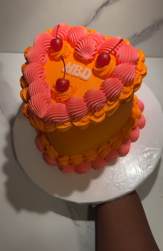 A two-tiered birthday cake decorated with yellow and pink frosting, topped with cherries and a "HBD" (Happy Birthday) sign, on a silver cake board.