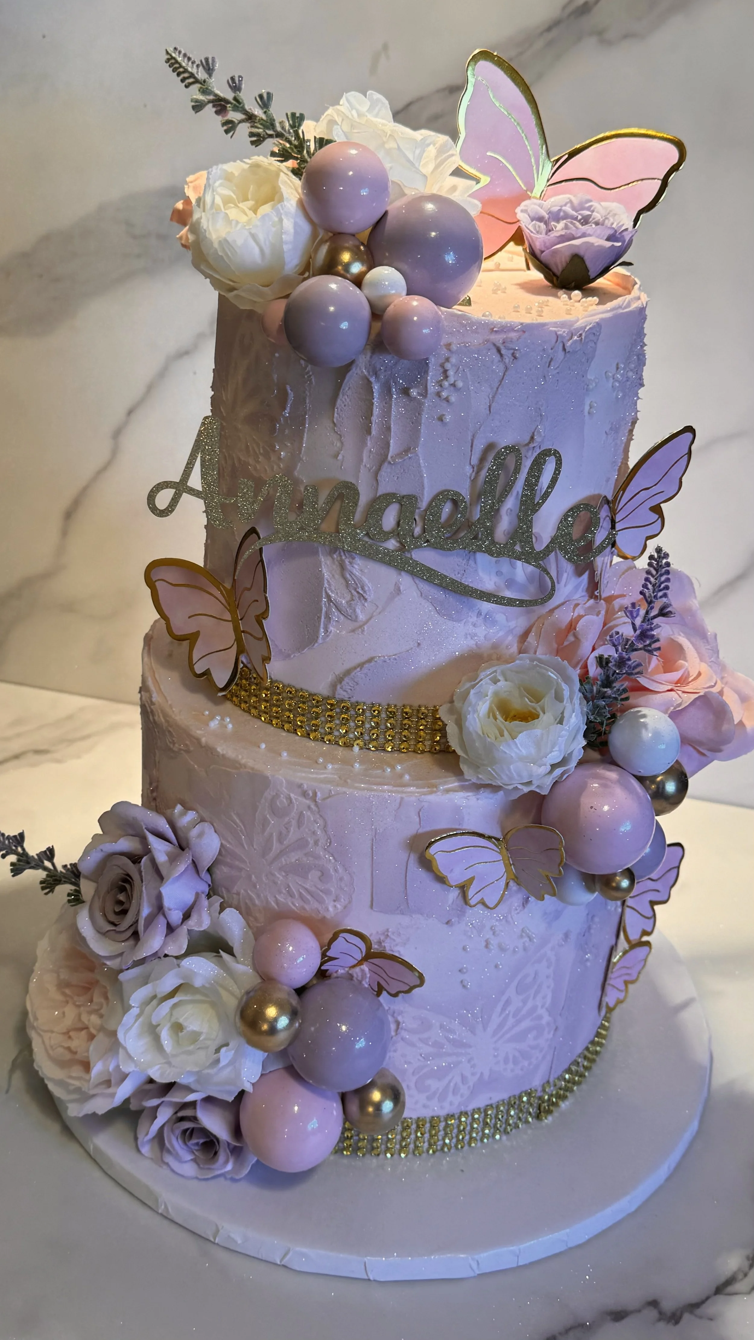 A two-tiered pink birthday cake decorated with flowers, butterflies, and pastel-colored balls, with a gold glitter sign that reads "Amelia".