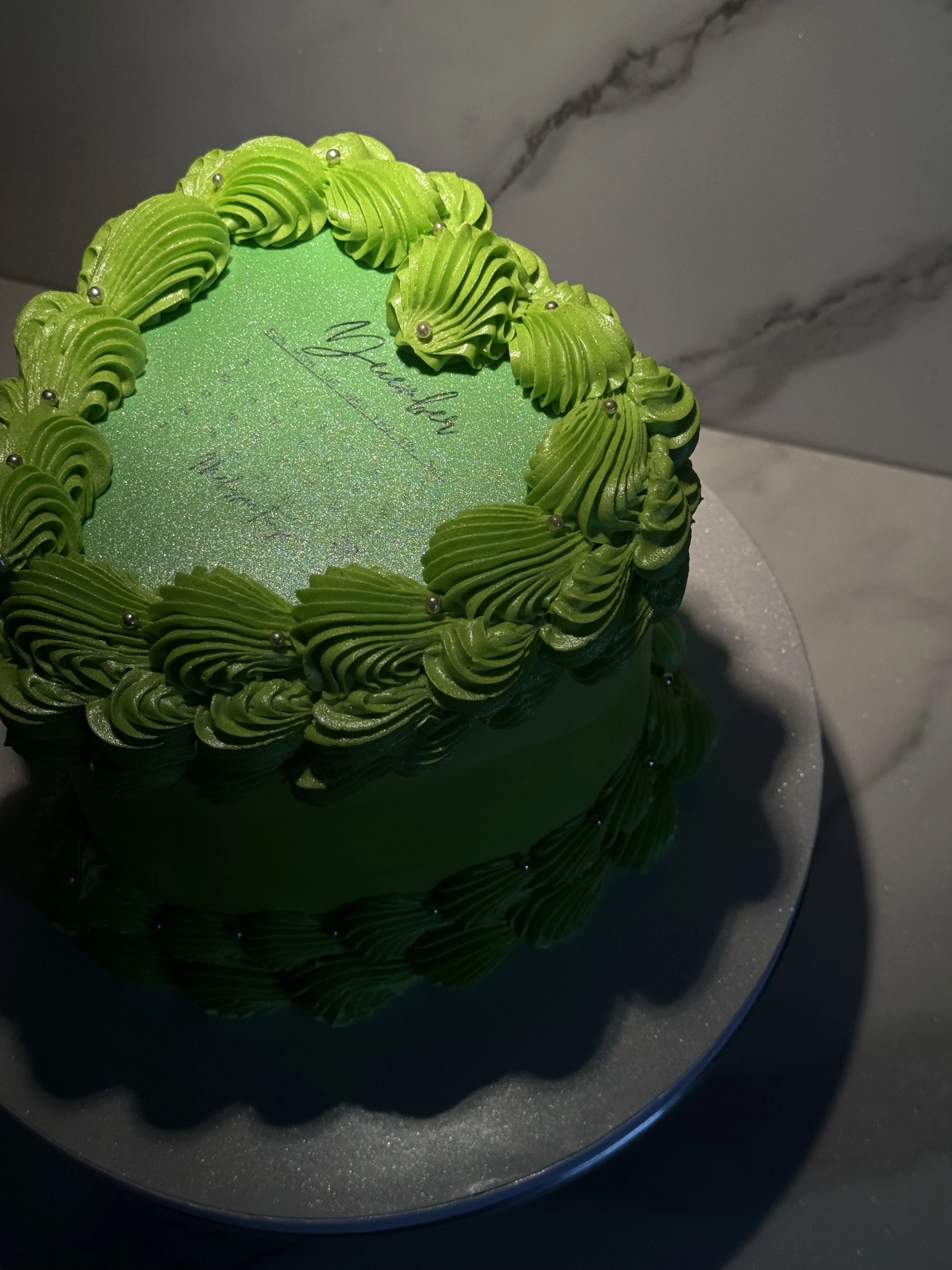 A round green cake with piped green frosting decorations around the top edge and a iridescent green top surface with a handwritten message, placed on a reflective silver cake board.