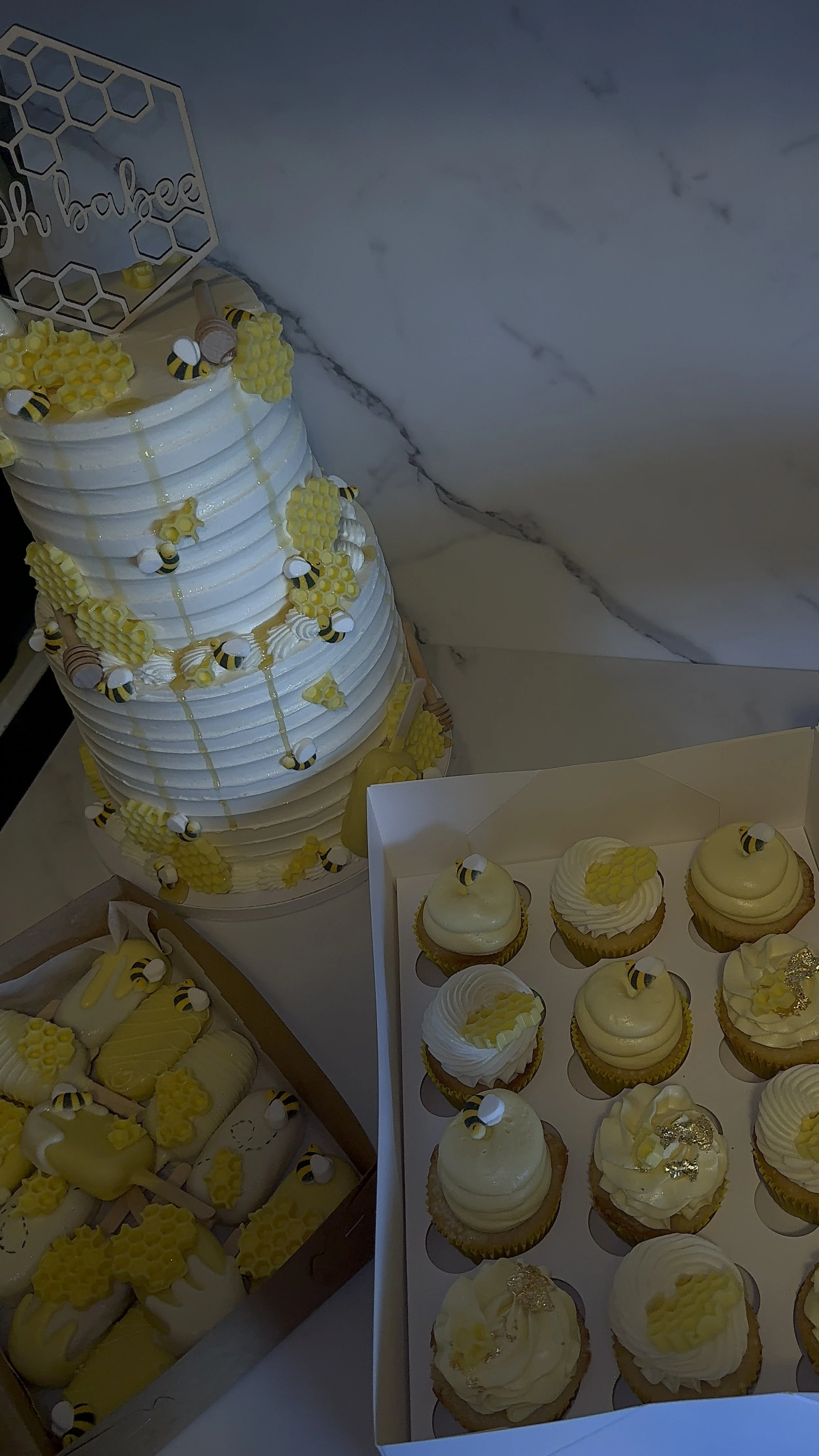 A tiered white cake decorated with small bee and honeycomb-shaped decorations, with additional cupcakes and honey-themed treats featuring bees and honeycomb patterns.
