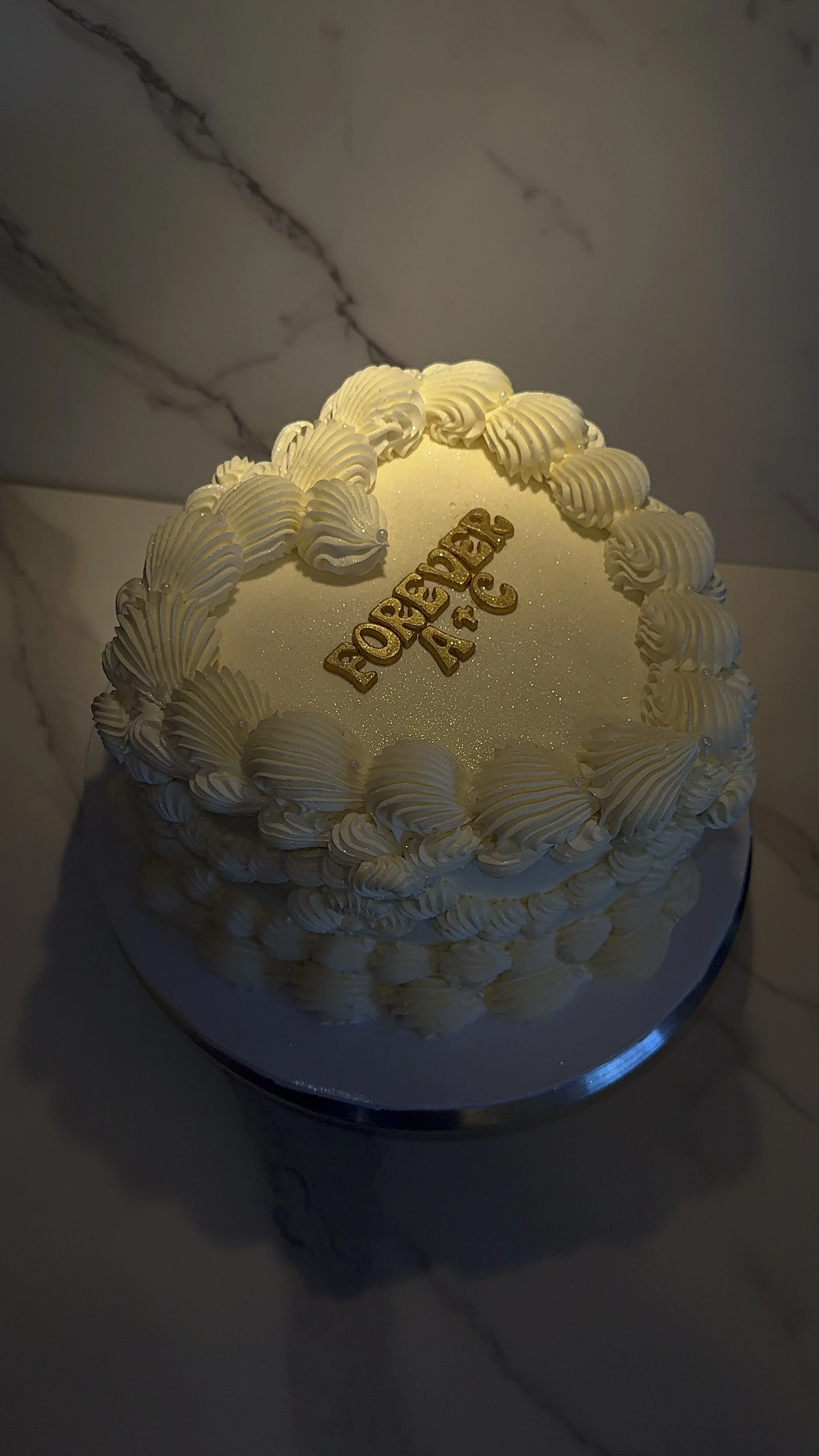 A white birthday cake with decorative piped frosting around the top edge and on the sides. The top has a glittery surface with a gold 'Forever & Always' cake topper. The cake is placed on a silver cake board on a marble surface.
