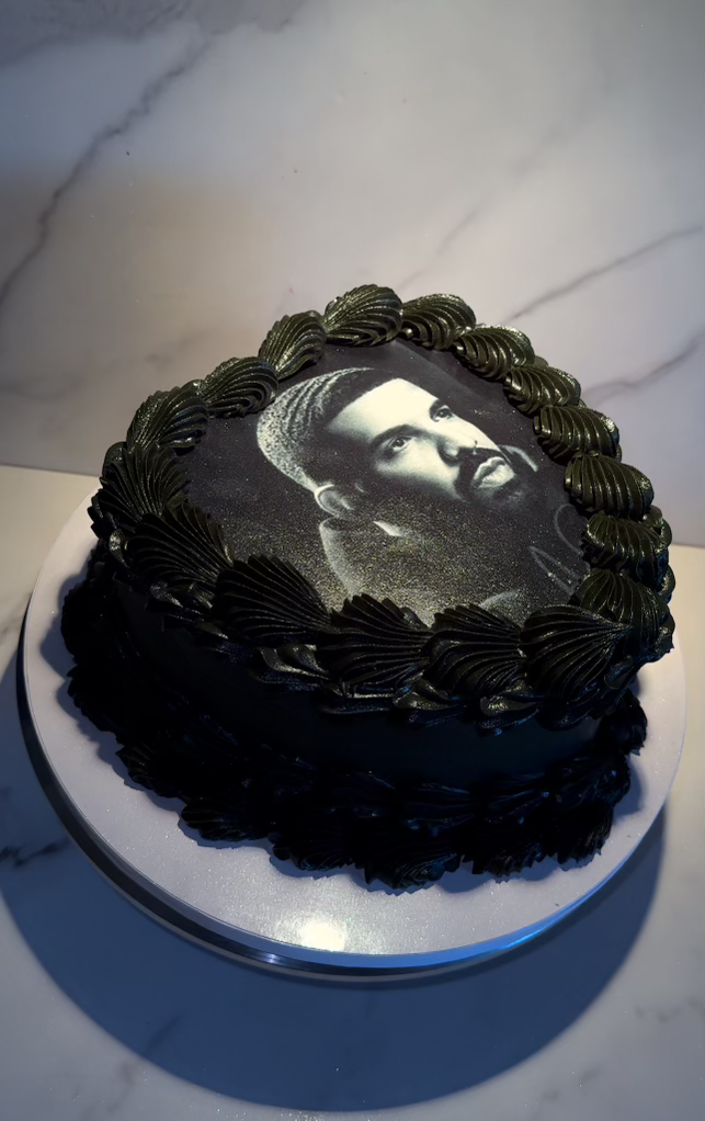 Black cake with decorative borders around an edible image of a man with a beard and mustache on top.
