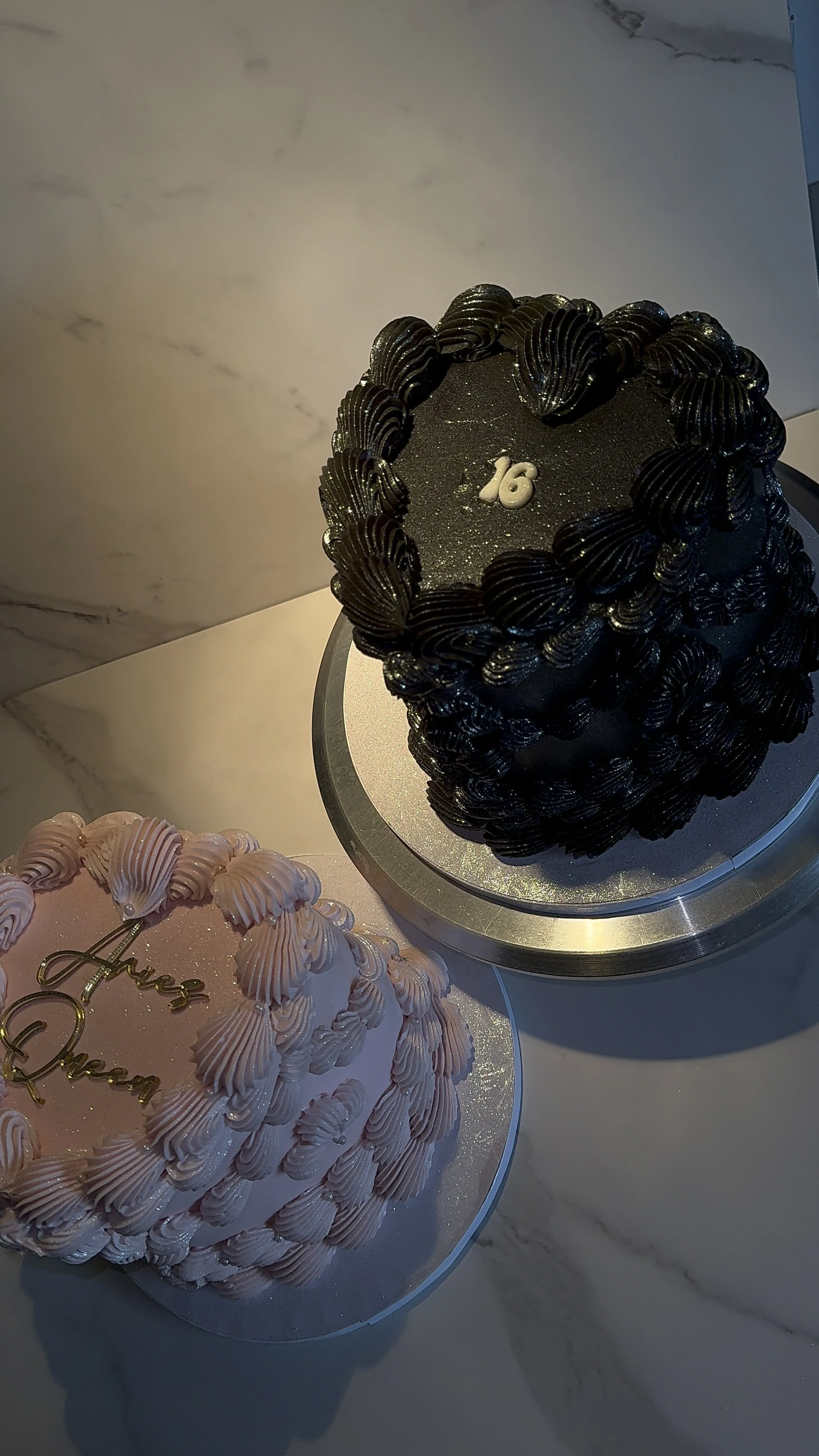 Two decorated cakes on a marble countertop, one black with a '16' decoration and ruffled piping, and one pink with pearls and 'Sweet Dream' writing.