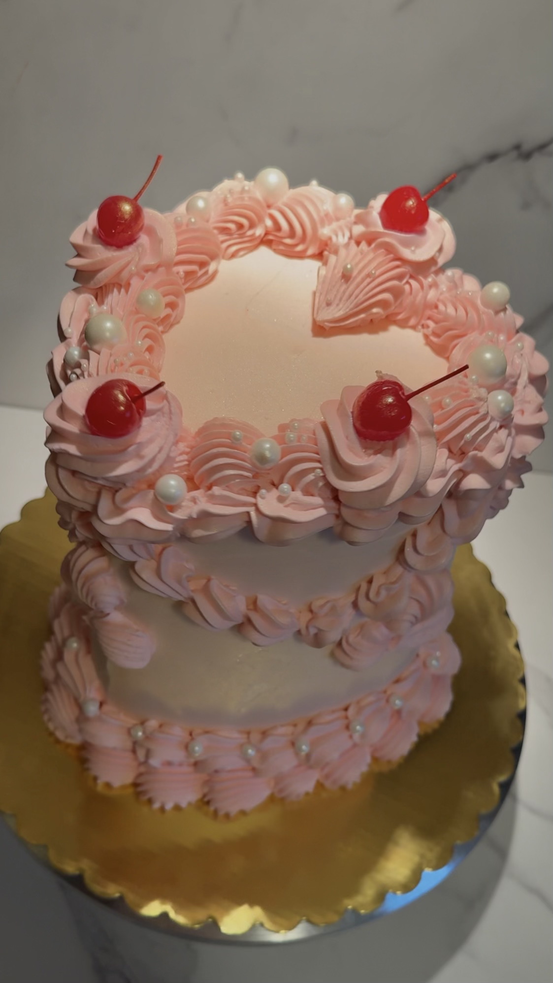 A two-tiered pink and white cake decorated with pink frosting swirls, white pearls, and red cherries on top.