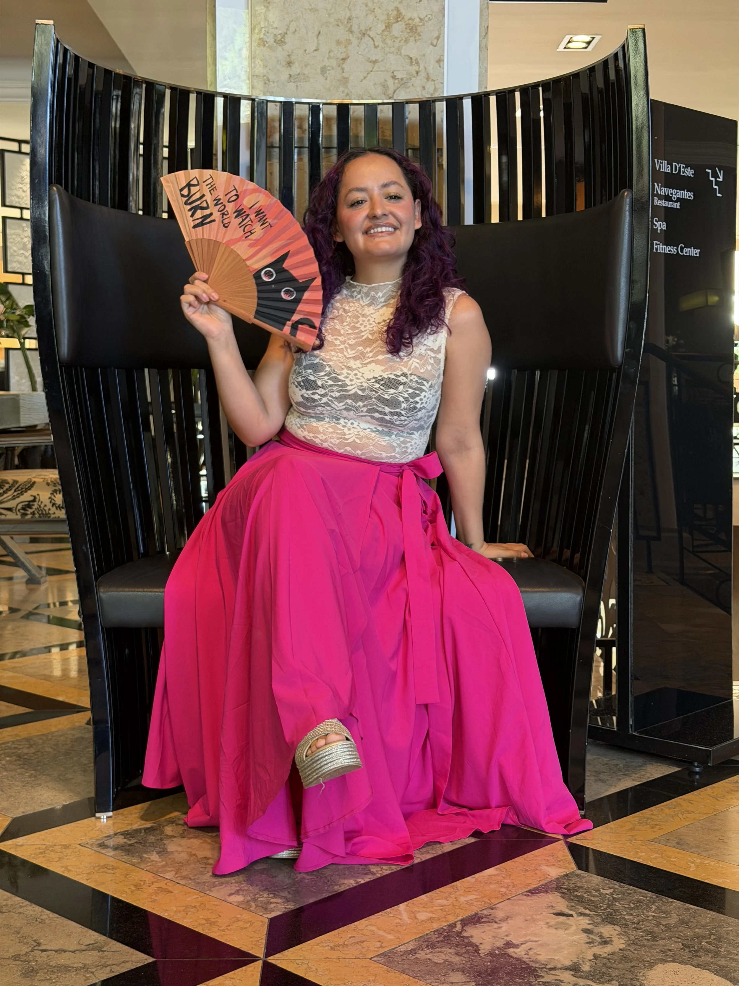 A woman with curly purple hair sitting on the floor, wearing a white lace top and a bright pink long skirt, holding a fan with the words "I want to be in the world" written on it, smiling at the camera. The background features a modern indoor space with black chairs, marble flooring, and directional signs.