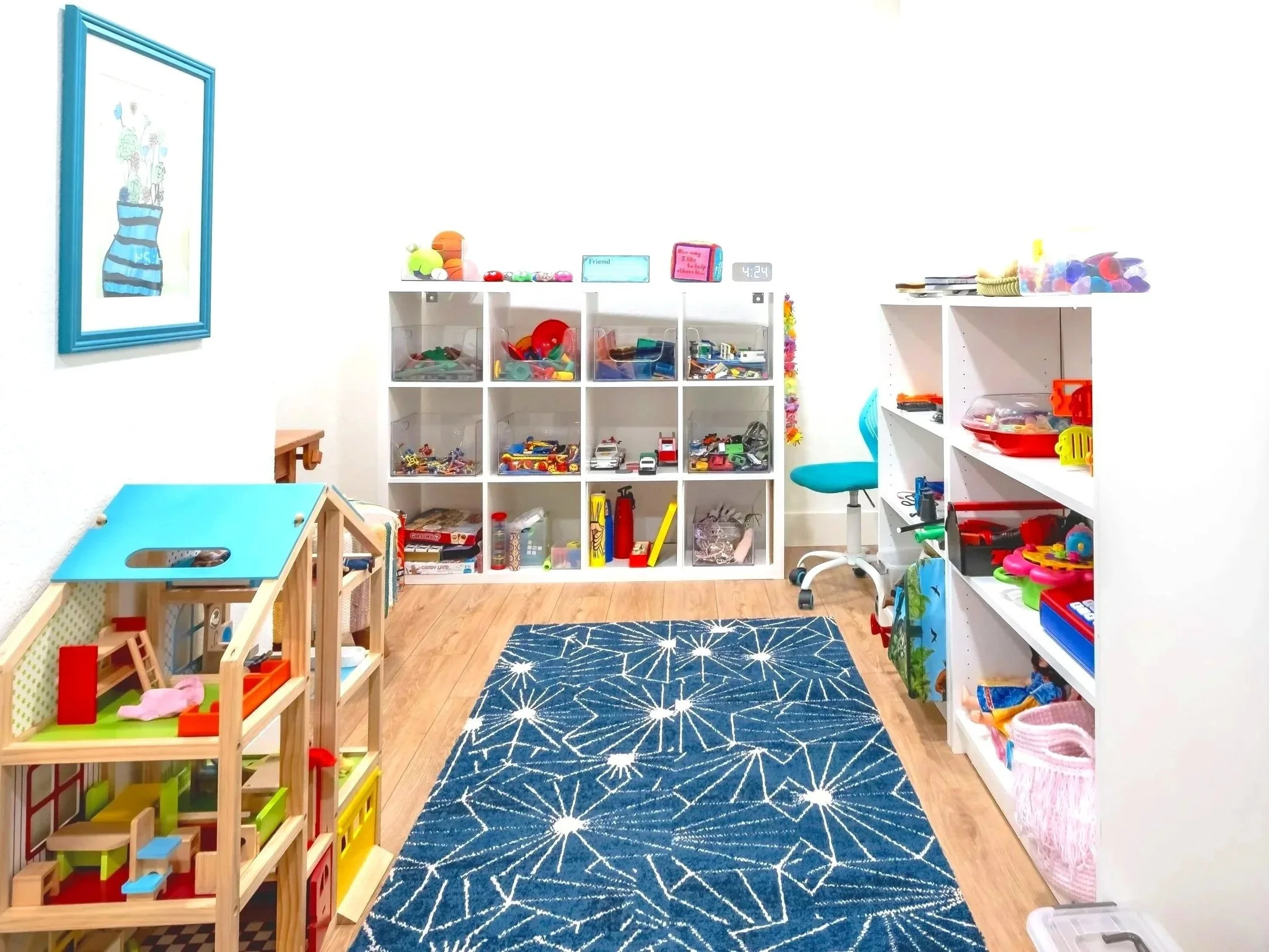 Toy storage with bins filled with toy cars, figurines, and other small toys. Small chairs and a play table on a blue patterned rug in a children's playroom.