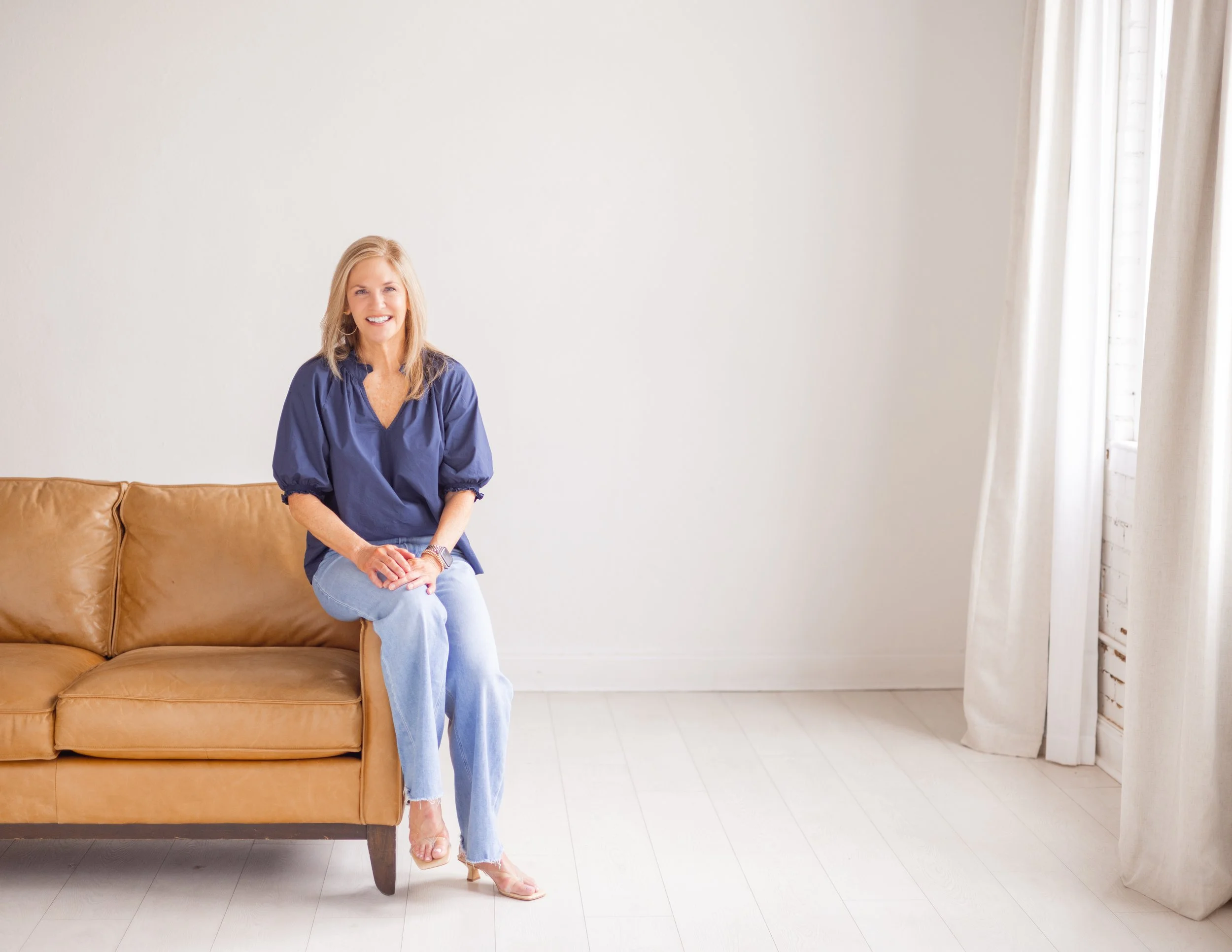 A woman with blonde hair and a blue blouse sitting on a tan leather sofa in a bright room with a white wall and curtains.