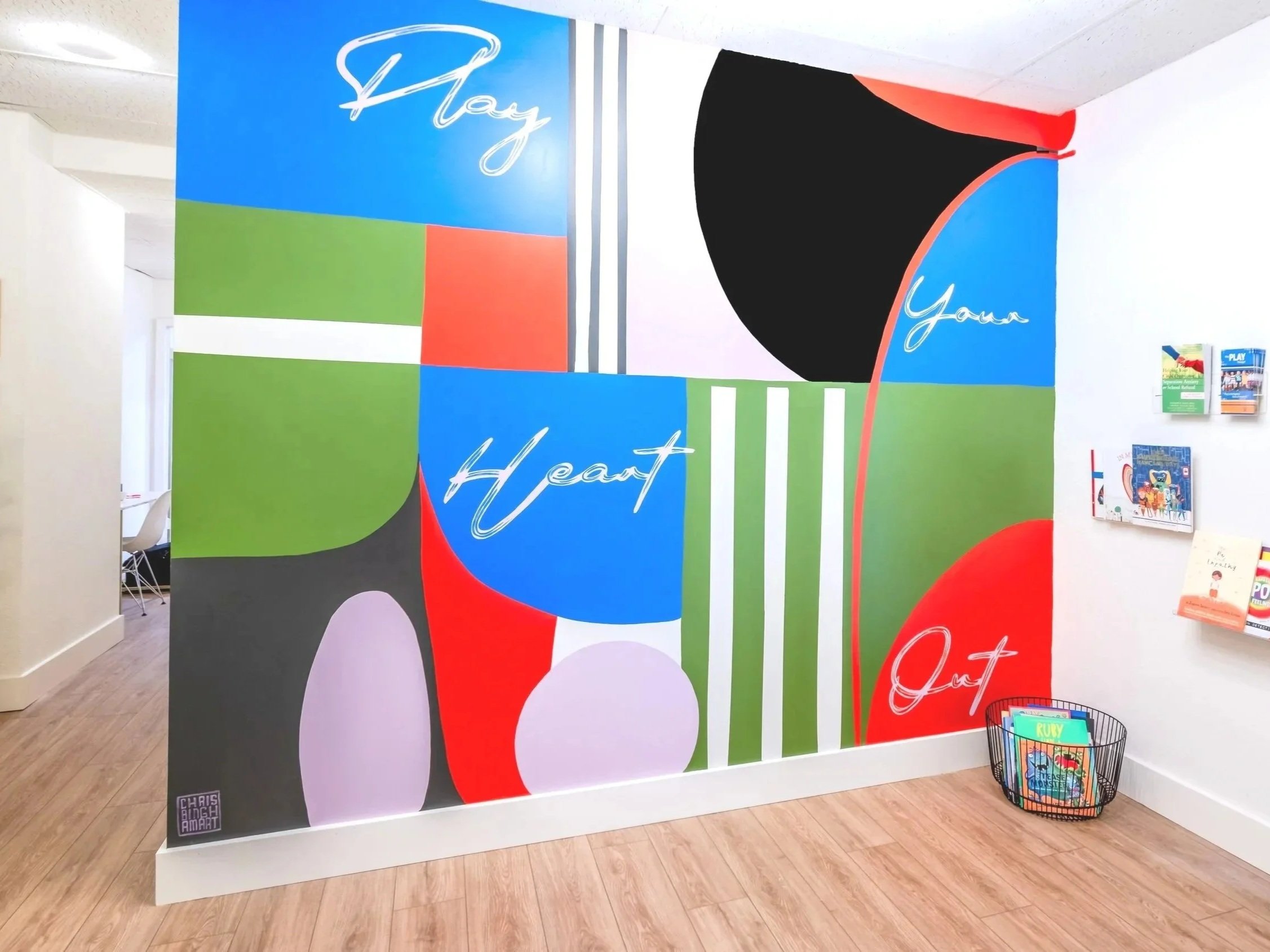 Colorful wall mural with abstract shapes and the words 'Hear Your Heart' written in cursive, situated in an indoor space with a wooden floor and some chairs and a wire basket with books nearby.