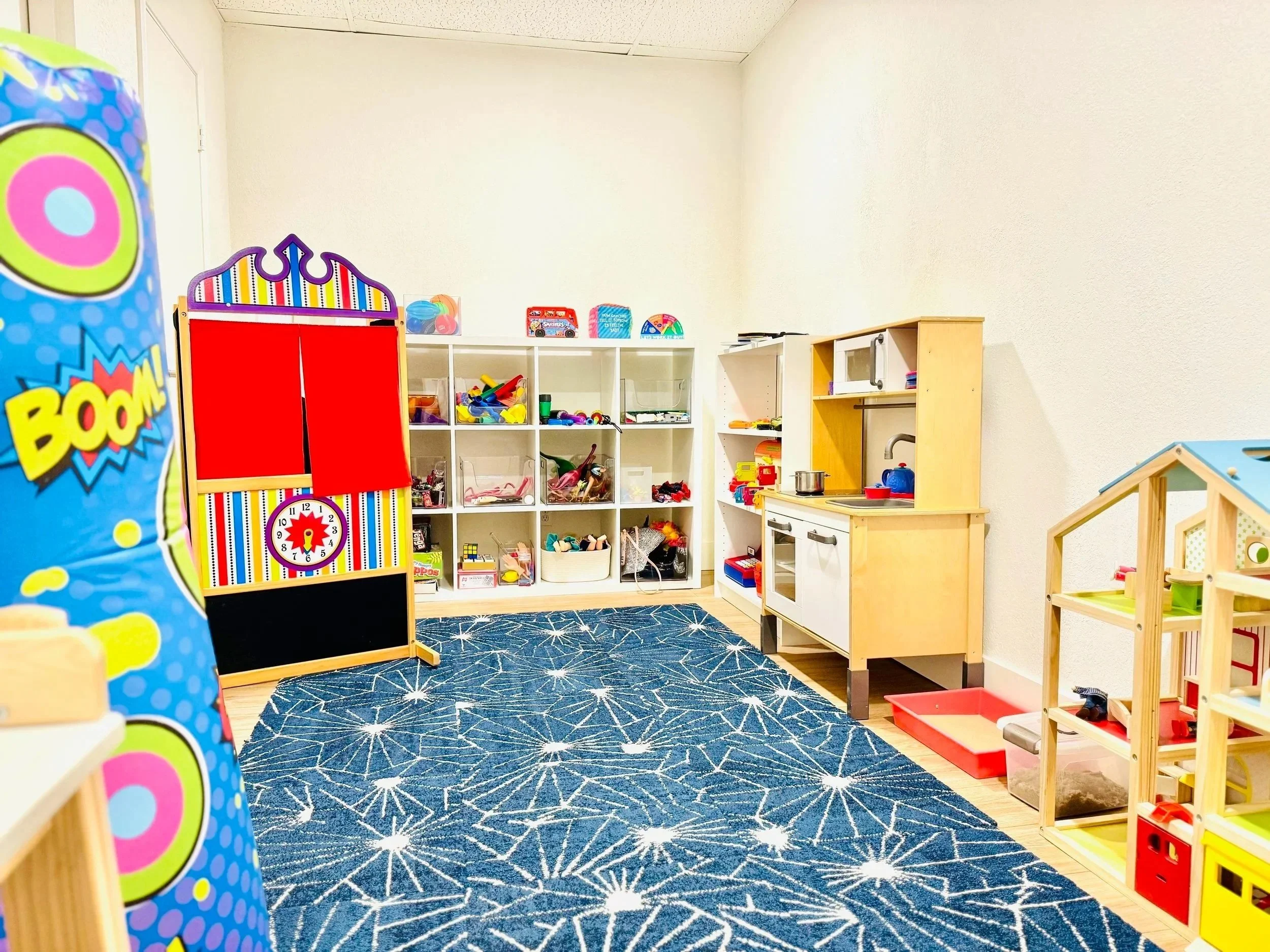 A corner of a children's playroom with shelves containing toys, a small toy kitchen, and a blue patterned rug on the wooden floor.