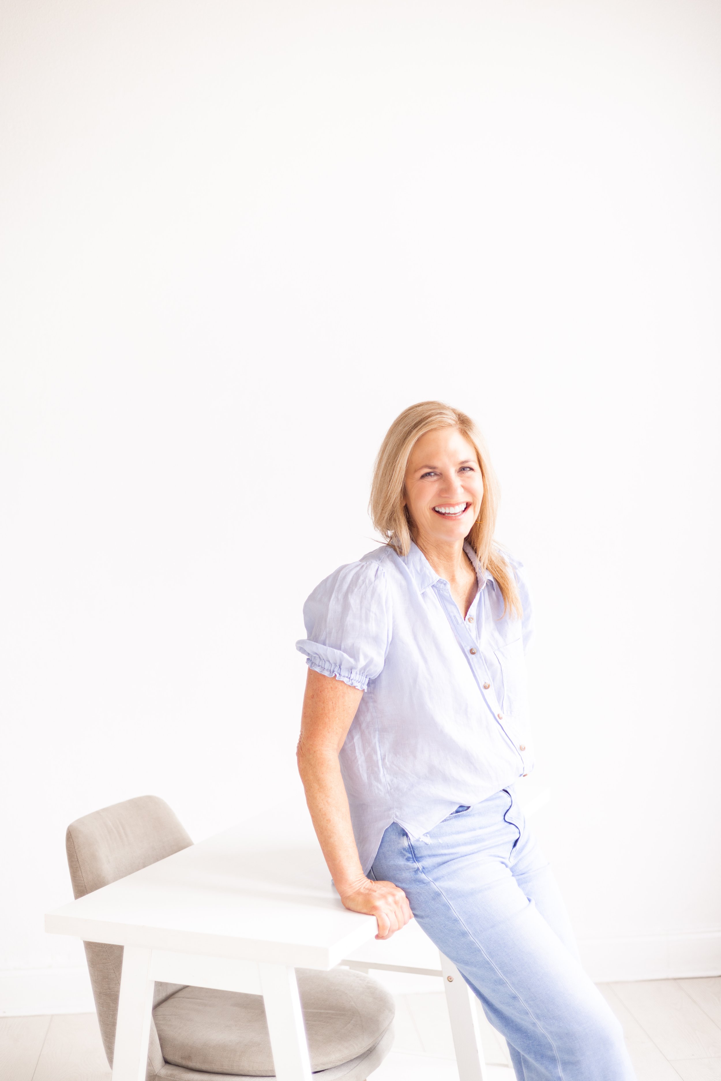 A smiling woman with blonde hair, wearing a light blue shirt and jeans, leaning on a white table in a bright, minimalistic room