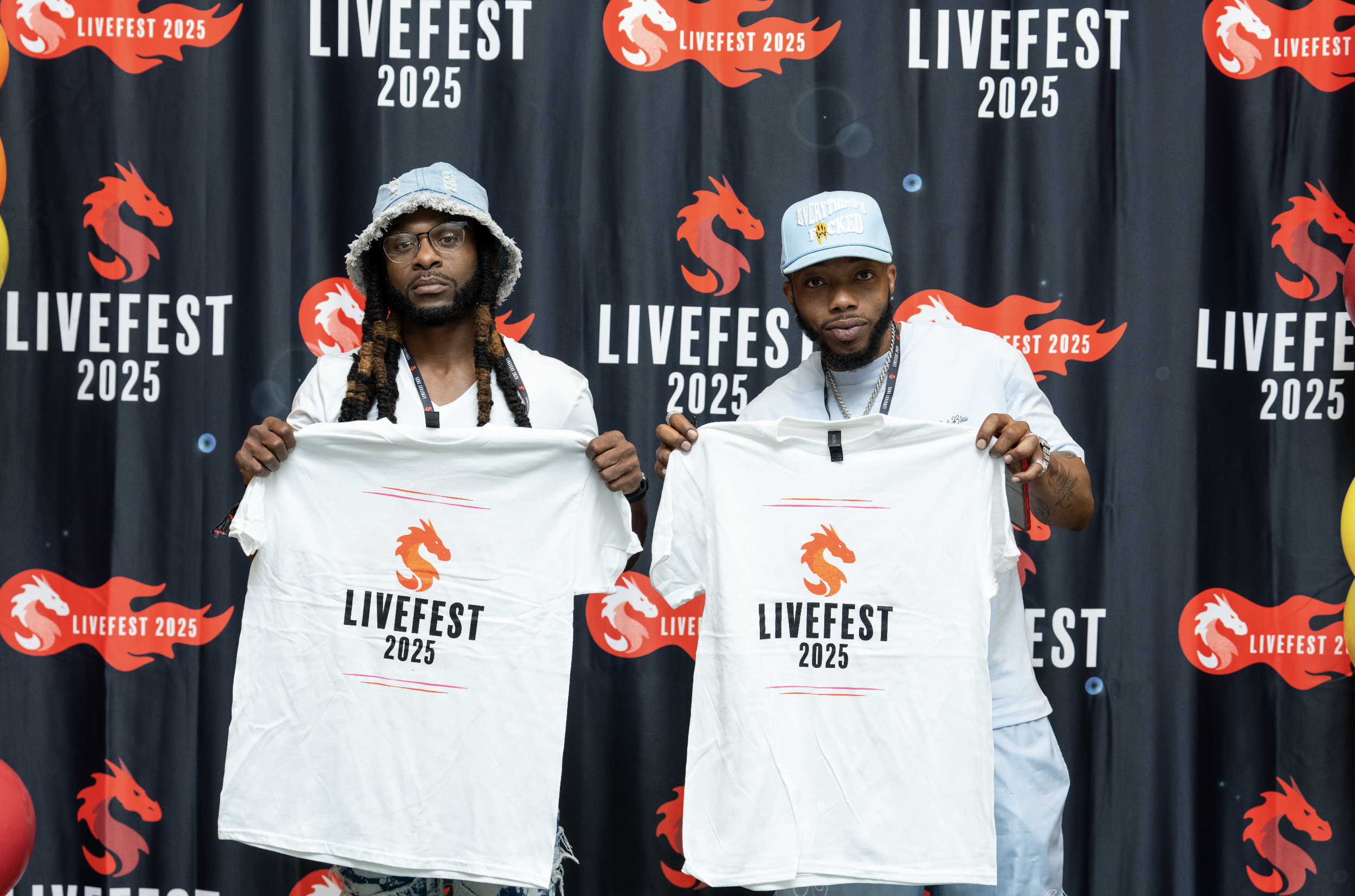 Two men standing in front of a black backdrop with red dragon logos and the words 'LIVEST 2025'. They are holding white T-shirts with the same logo and words. One man wearing a bucket hat with a hoodie and glasses, the other wearing a baseball cap an