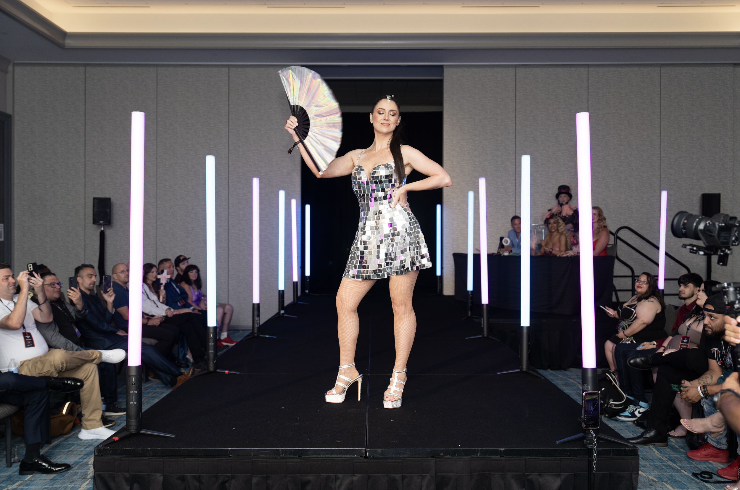 A woman in a reflective, mirror-like dress and high heels walking on a runway, holding a fan, with an audience and colorful vertical light poles on either side.