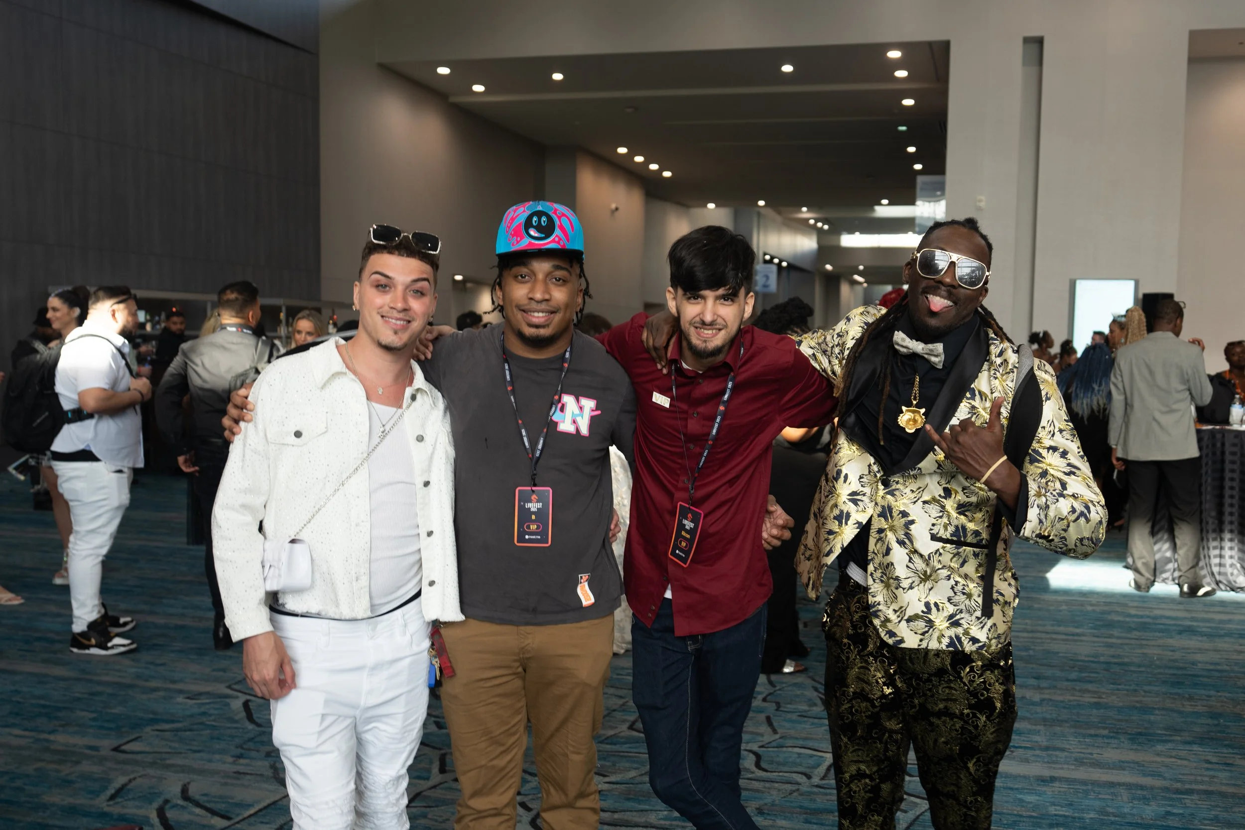 Four men posing together at an indoor event, smiling and with their arms around each other. The man on the far left is wearing a white denim jacket and pants, the second man is in a gray t-shirt with a colorful helmet, the third man is in a maroon sh