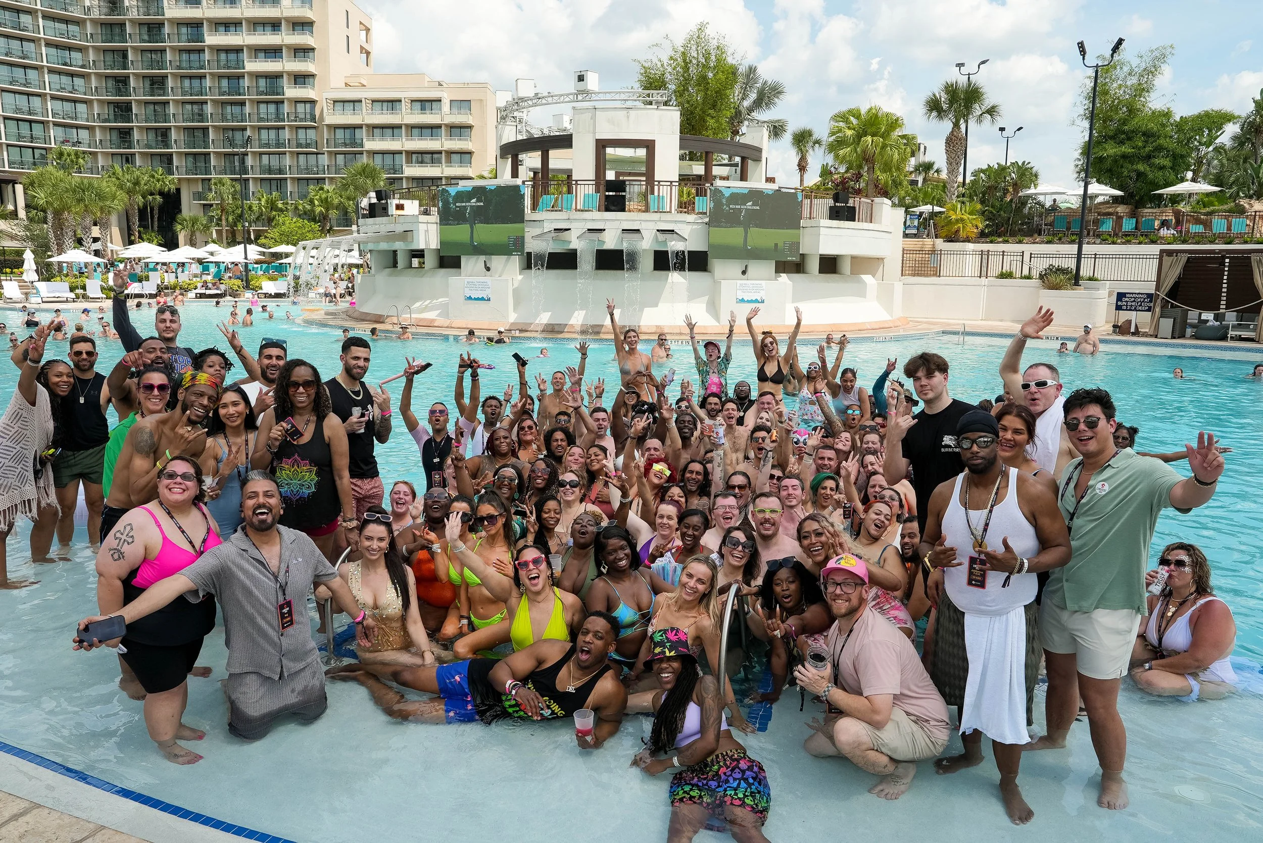 A large group of people in swimsuits and casual clothing standing in a swimming pool at a resort, smiling, waving, and making peace signs. In the background, there are palm trees, umbrellas, and a multi-story building, with a poolside bar or stage ar
