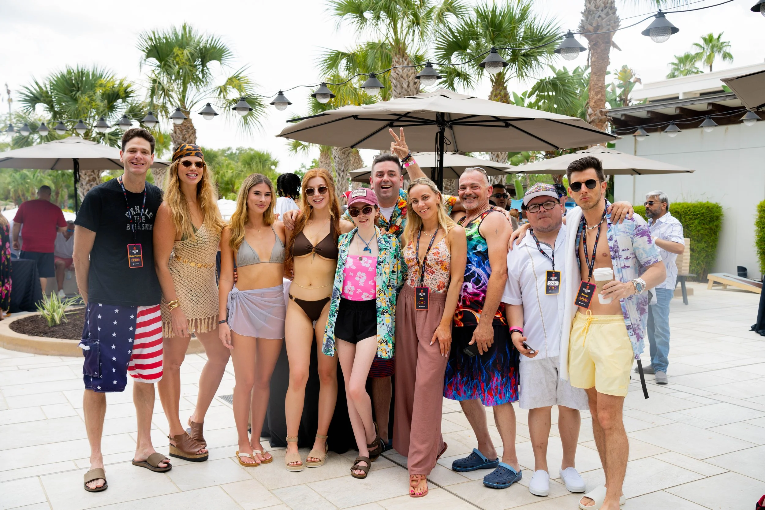 Group of people in summer casual clothing and swimsuits, standing outdoors at a poolside party with palm trees and umbrellas in the background.