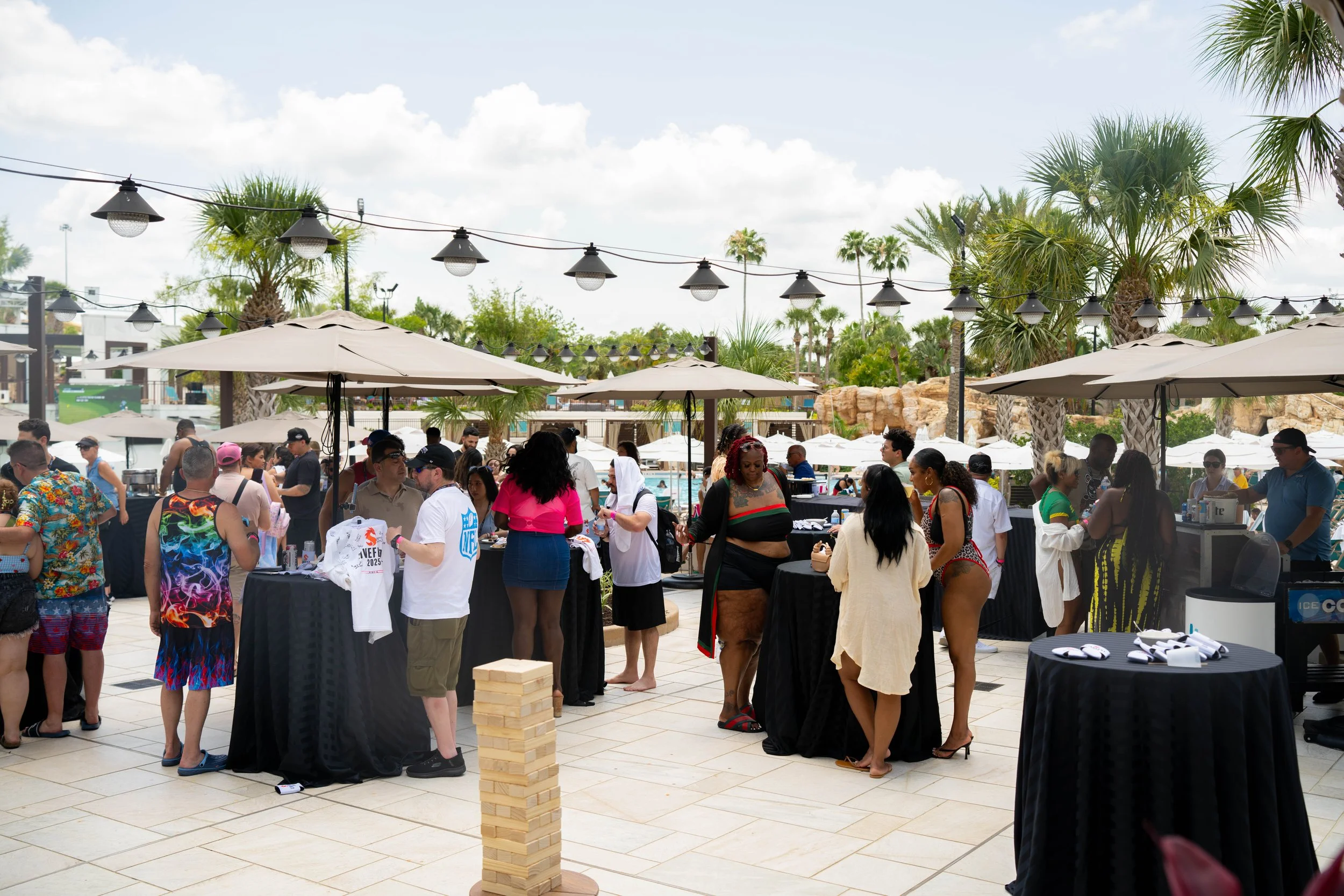 Crowd of people at an outdoor event or pool party, standing around tall tables with black tablecloths, under umbrellas and string lights, with palm trees and a pool in the background on a sunny day.