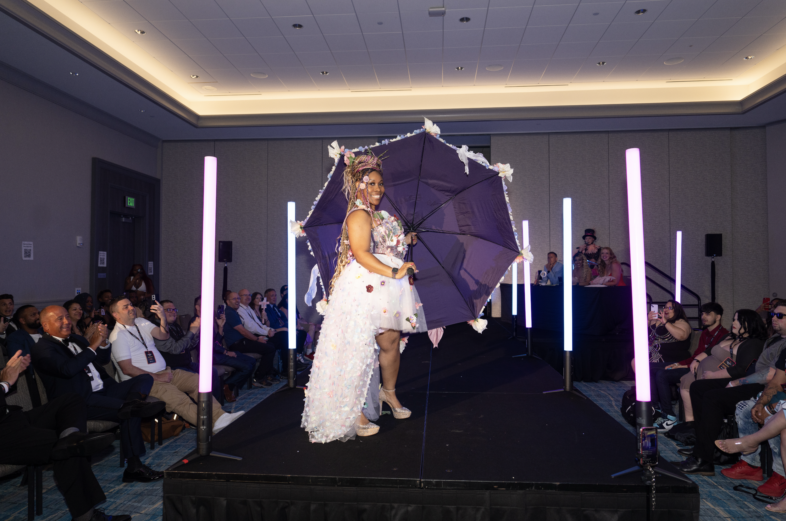 A woman standing on a runway stage is holding a giant purple umbrella decorated with flowers. She is smiling and wearing a white dress with floral accents and platform shoes. The audience is clapping and taking photos, and colorful lights are ambient