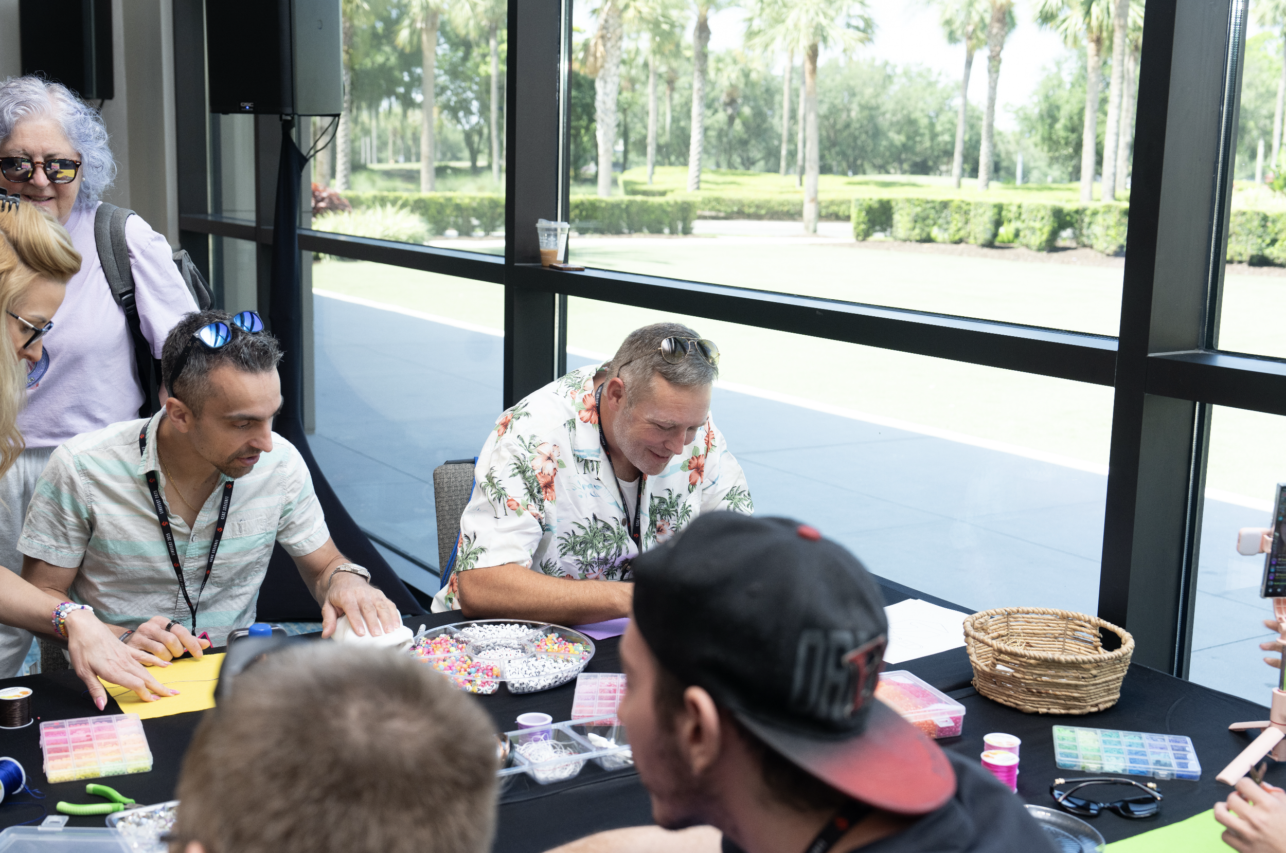 Group of people decorating craft items at a table in a bright room with large windows and a view of a park outside.
