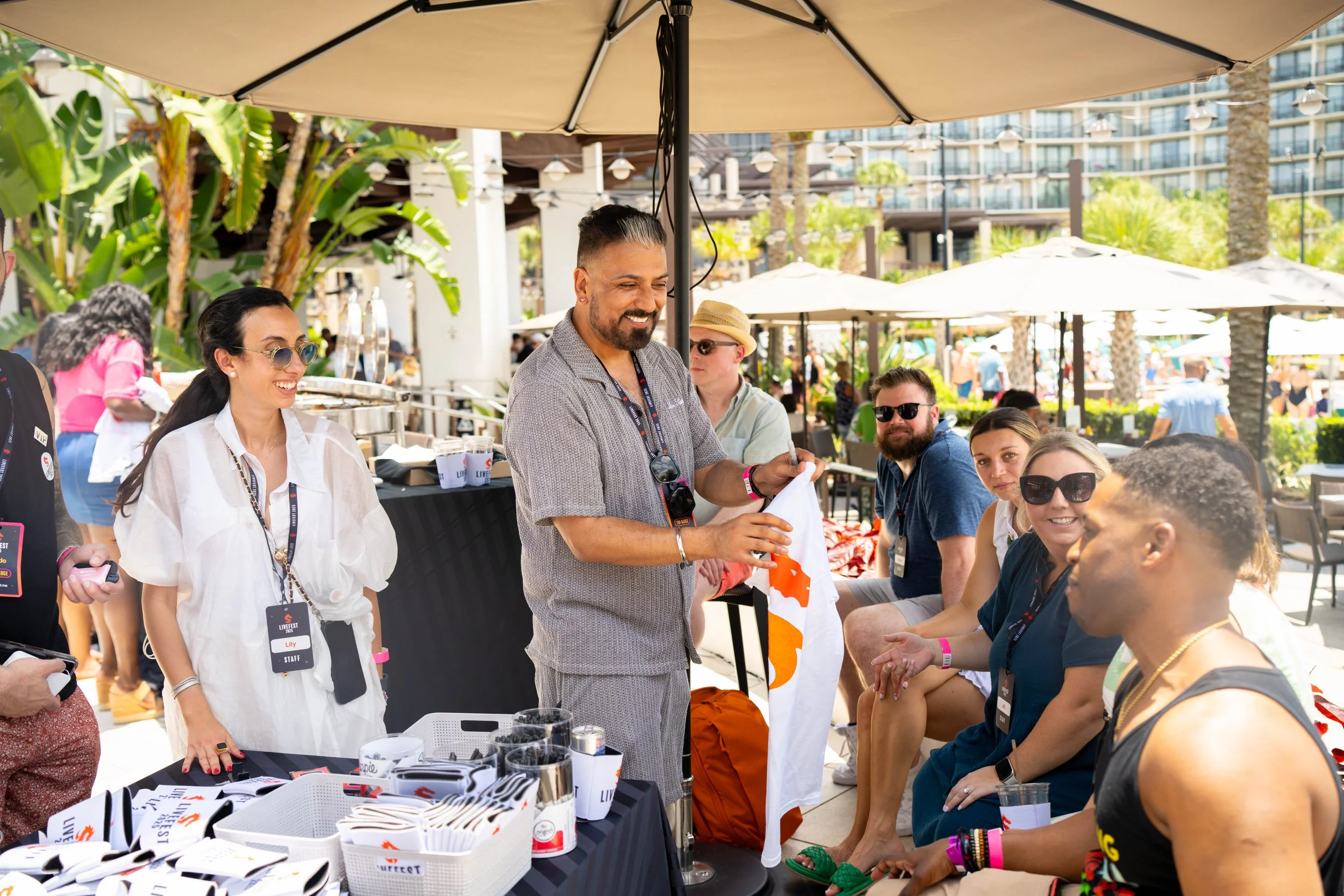 Group of people at outdoor event under umbrellas, some sitting, some standing, with vendor tables and background of palm trees and buildings.