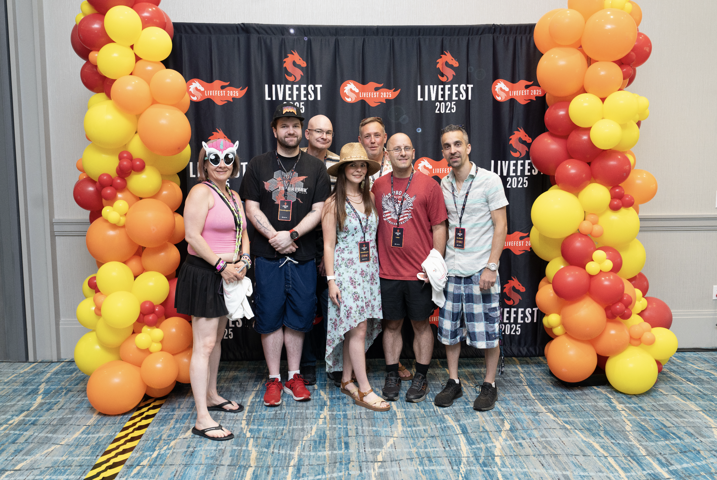 A group of seven people standing in front of a black backdrop decorated with red and yellow balloons and the Livfest 2025 logo. The group includes four men and three women, with some smiling and others looking at the camera. One woman is wearing a un
