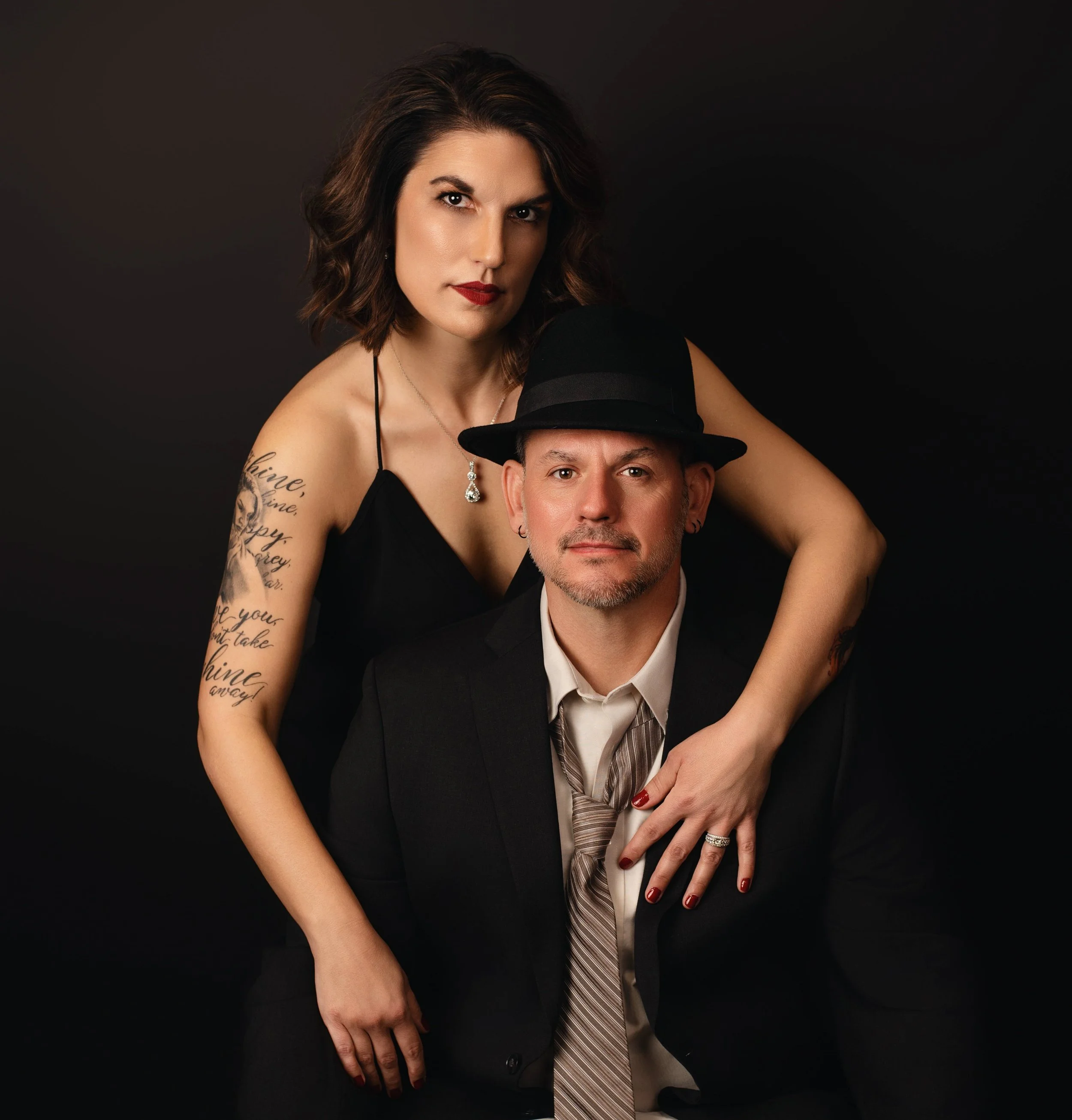 A woman with a tattoo on her arm stands behind a man wearing a black suit, white shirt, and black hat.