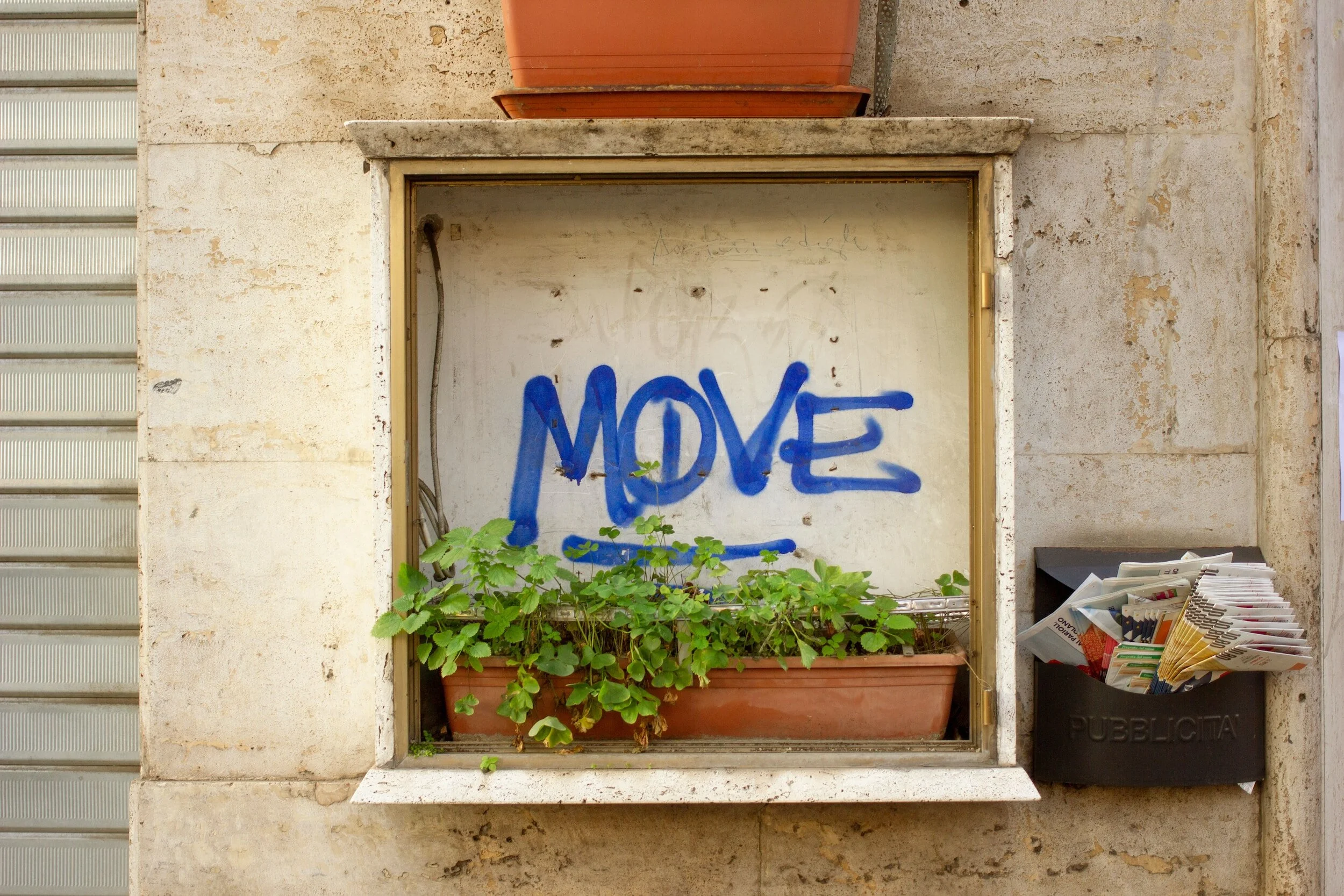Urban scene with a window box containing green leafy plants and graffiti text "MOVE" in blue on the wall. A black mailbox filled with brochures and newspapers labeled "PUBBLICITÀ" is on the right.