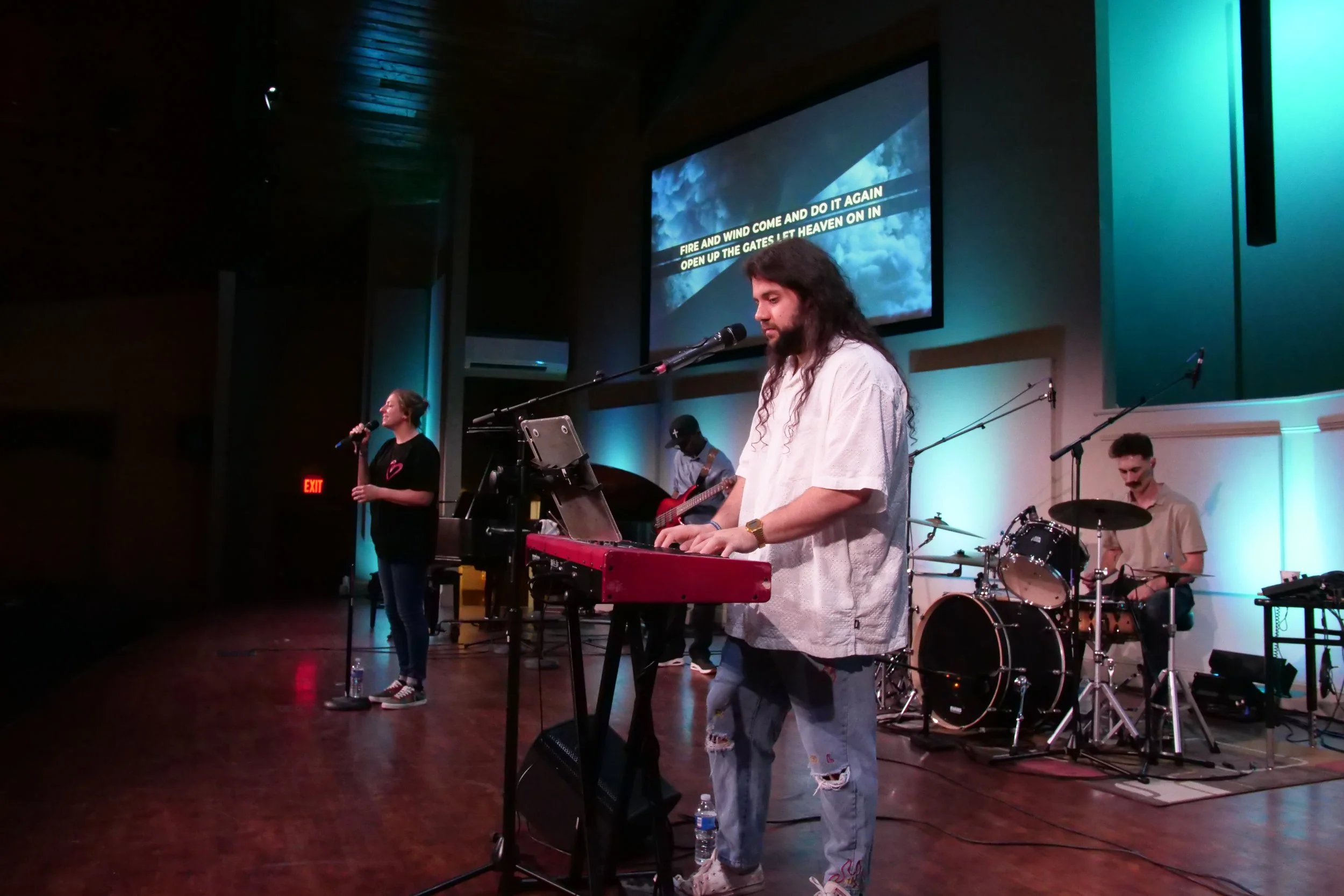 A band performing on stage with a singer holding a microphone and a keyboard player playing a red keyboard. A bass guitarist and a drummer are also visible. Large screen in the background displays lyrics.