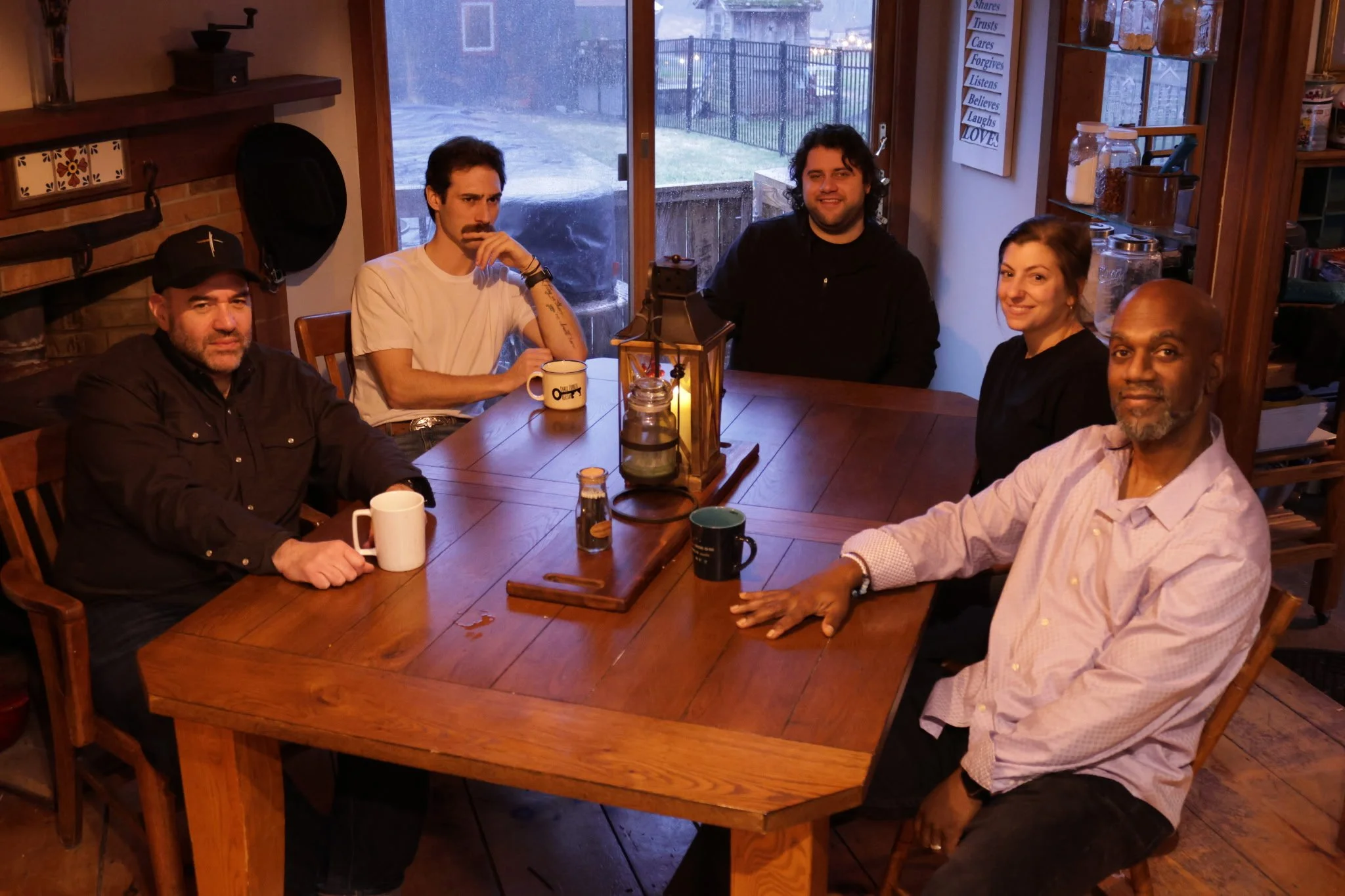 Five people sitting around a wooden dining table in a cozy kitchen with a window overlooking a backyard. The table has a lantern, a candle, and several mugs. The group appears relaxed and smiling, with one person looking serious.