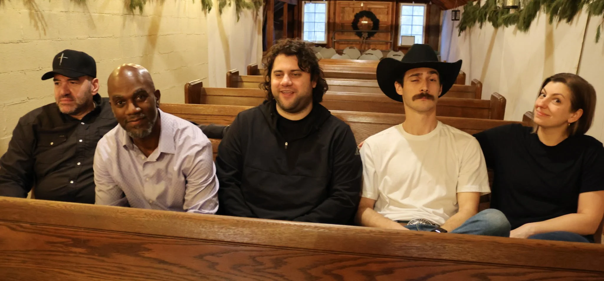 Five people sitting on wooden church pews in a rustic room with Christmas decorations. From left to right: a man wearing a black cap, a man with a bald head and beard in a white shirt, a man with curly hair and a black jacket, a man with a mustache w