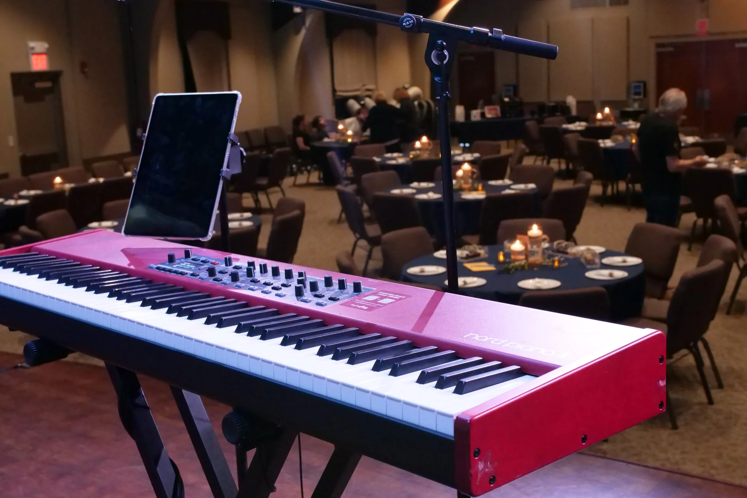 Red keyboard on a stand in a dimly lit banquet hall with round tables, candles, and people in the background.