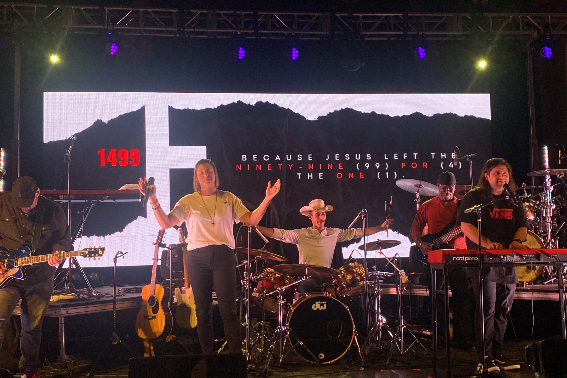 A band performing on stage with a large screen behind them displaying a biblical reference. The screen shows a torn paper design with the number '1499' in red on the left and text in white and red reading, 'Because Jesus left the ninety-nine (99) for the one (1).' The band includes a female singer with a microphone, a keyboard player, a drummer, a guitarist, and a keyboardist. The stage is lit with colorful lights, and the musicians appear engaged in their performance.