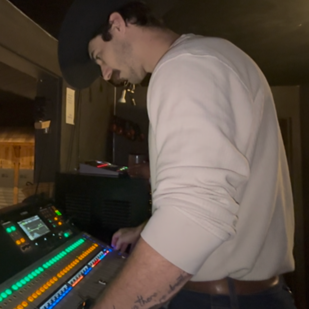 A man with dark hair and a black cap is working on a digital mixing console with colorful lights, in a dimly lit room.