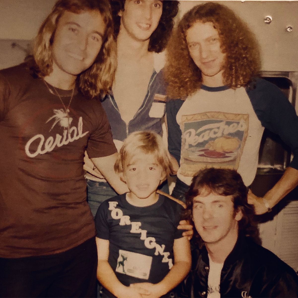 Group of five people, three women and two children, posing together in a casual setting. The women have curly hair and are wearing casual shirts, one with a 'Capital' logo and the other with a 'Pratt' logo. The children are a boy with blond hair and a girl with brown hair. All are smiling or making playful faces.