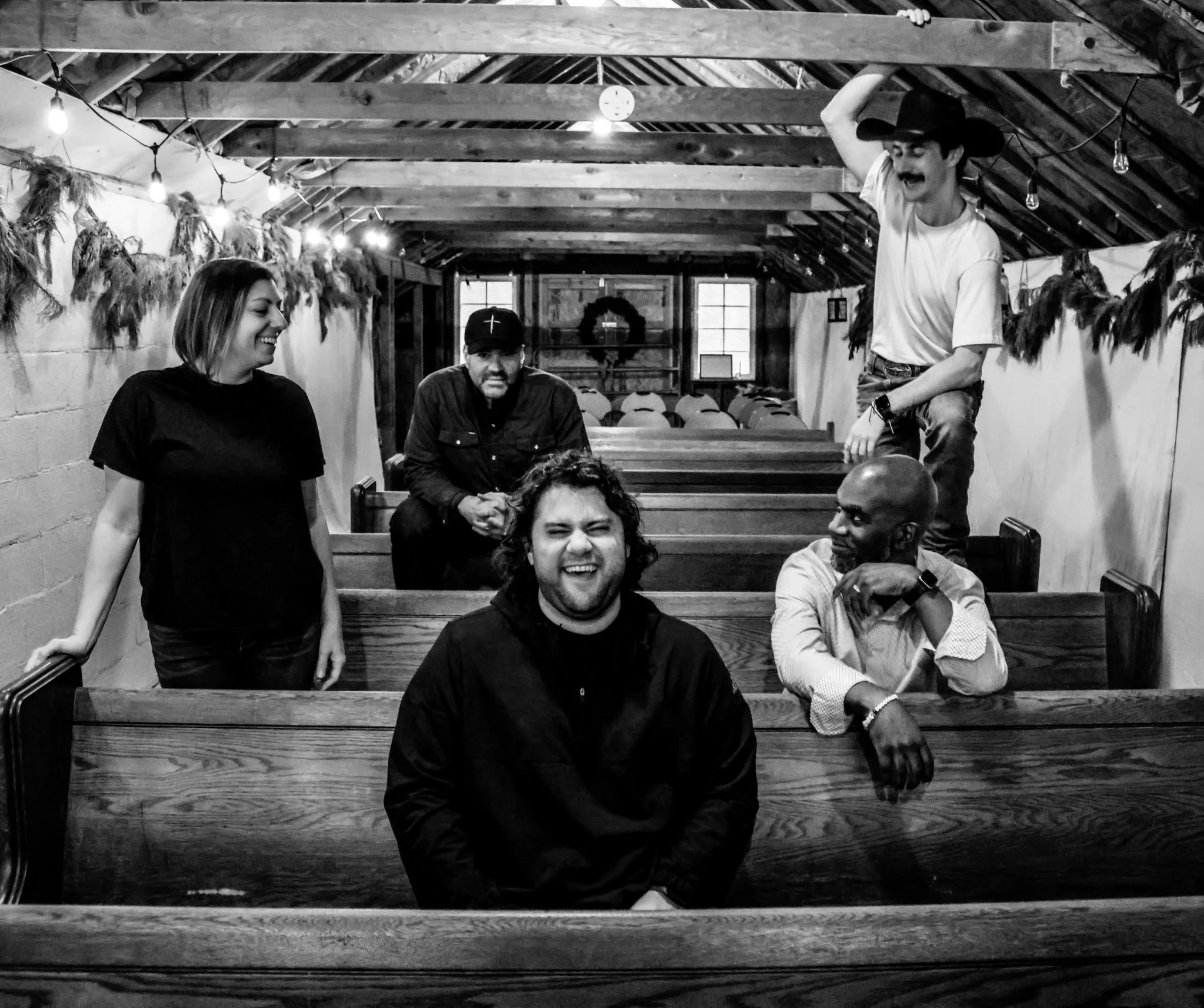 A black and white photo of 1499's five band members gathered inside a rustic, wooden barn decorated for Christmas with hanging lights and dried branches. They are smiling and laughing, enjoying each other's company.